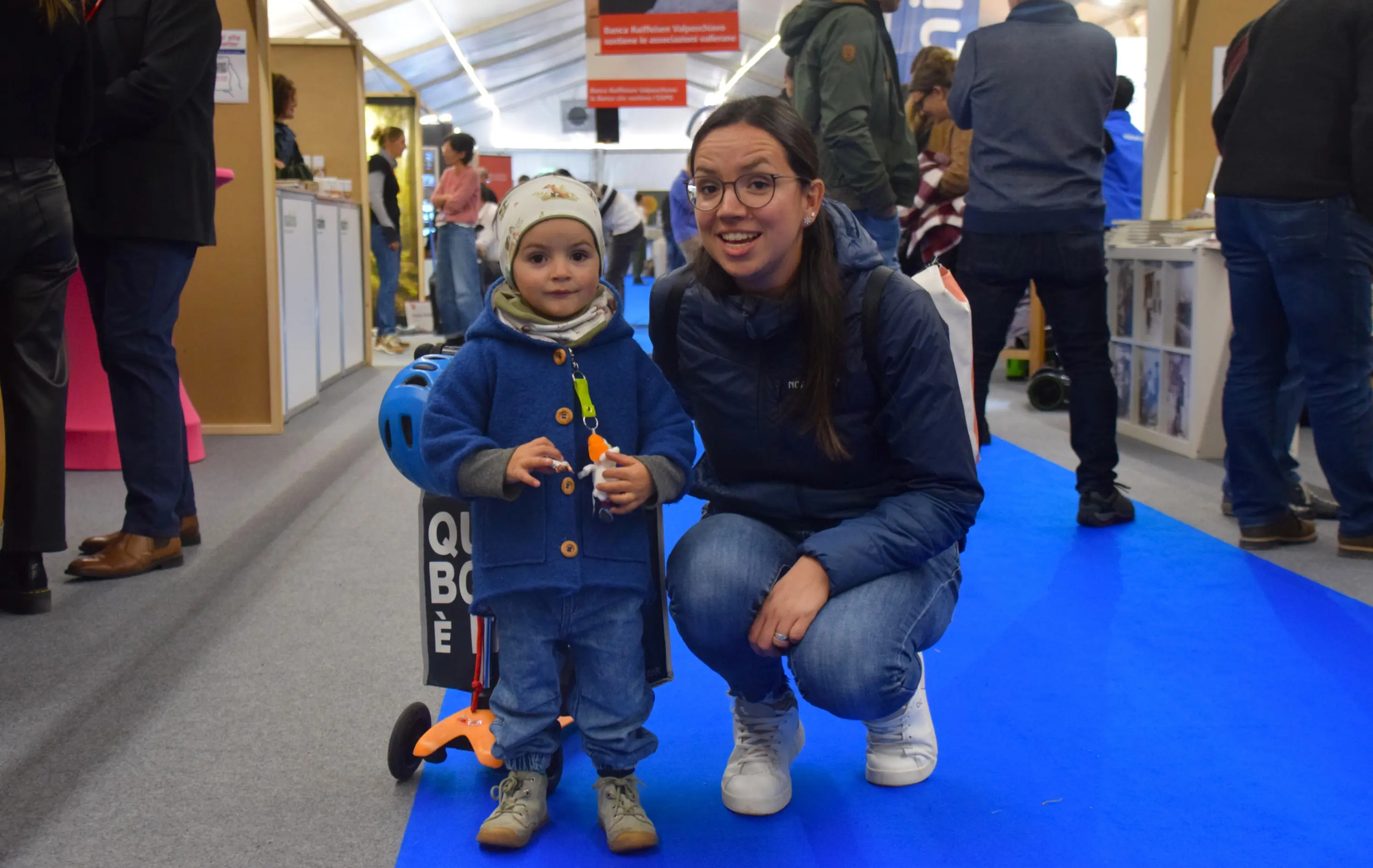 Un bambino in giacca blu con un berretto gioca con un carrello, mentre una donna sorridente in giacca blu si accovaccia accanto a lui. Un ambiente di fiera o evento con persone sullo sfondo.