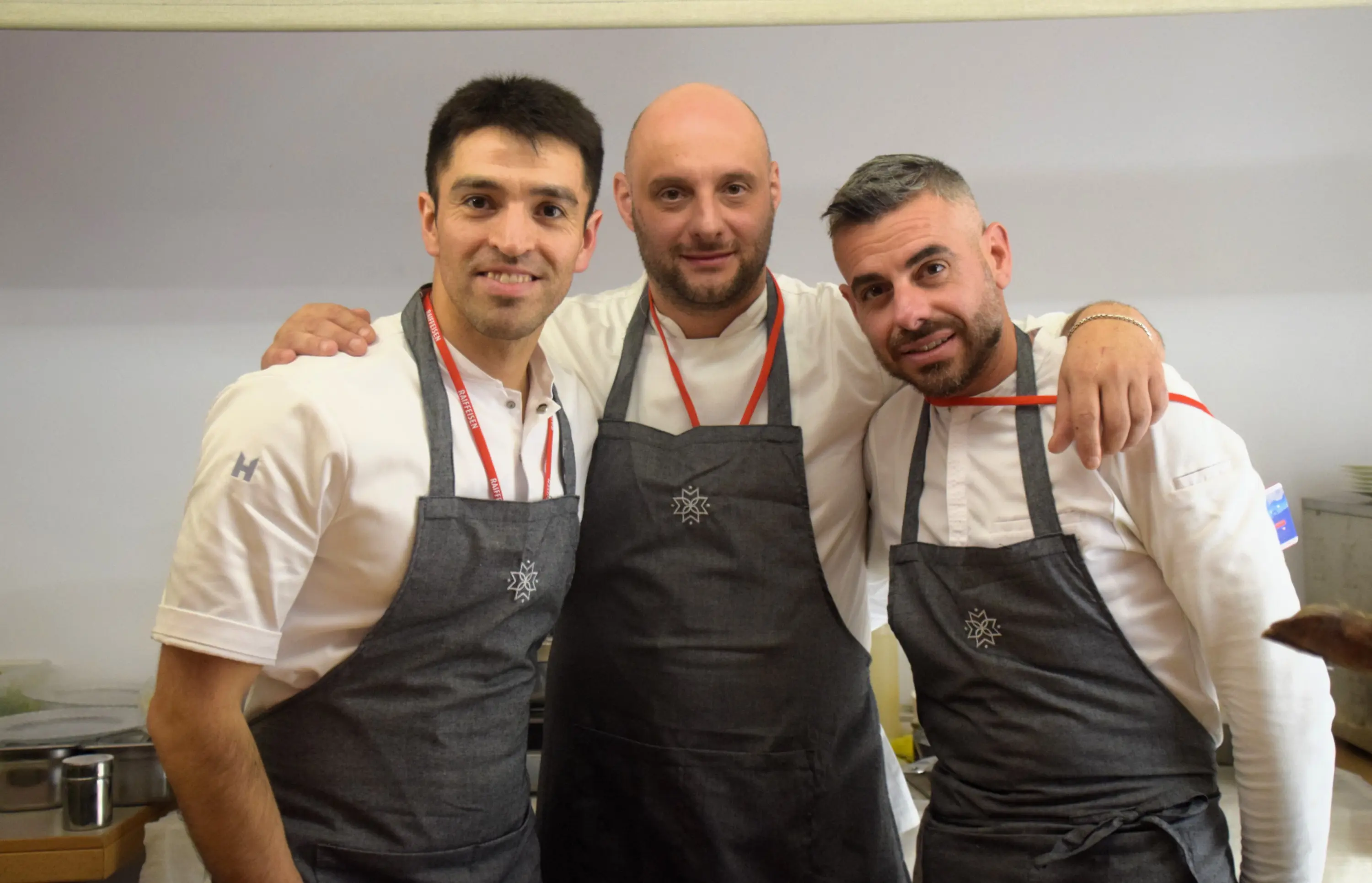 Tre chef sorridenti in uniformi da cucina grigie, con grembiuli e medaglie rosse, si abbracciano in un ambiente di cucina.