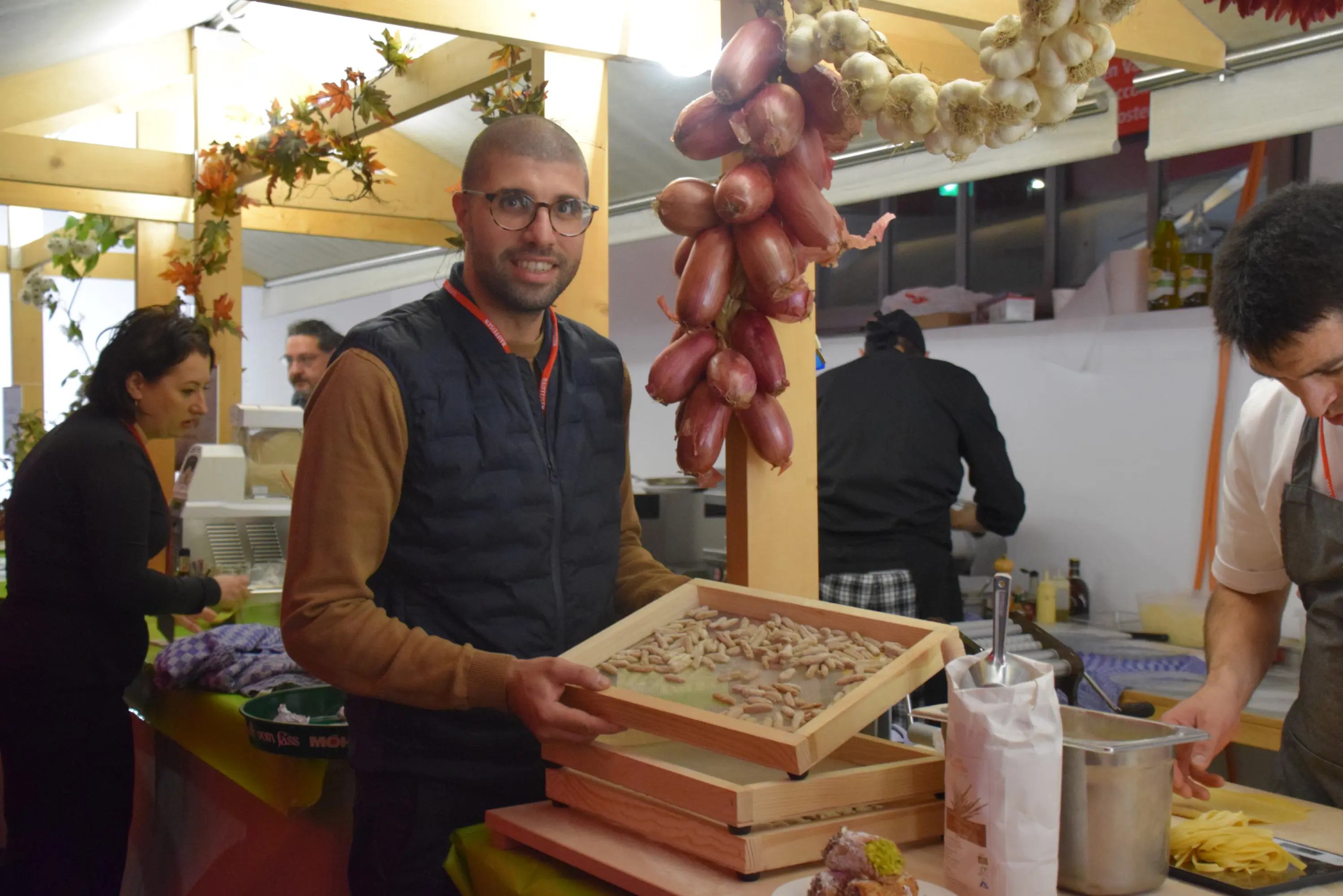Un uomo sorride mentre tiene un vassoio di ingredienti, circondato da prodotti freschi e uno stand gastronomico. Sullo sfondo, altre persone sono impegnate nella preparazione di cibo.
