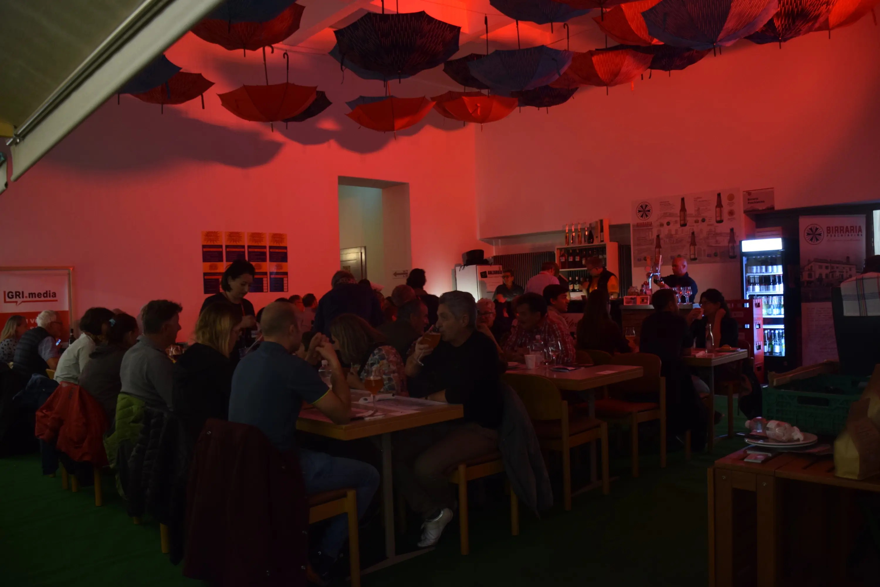 Un locale affollato con persone sedute a tavoli, luce rossa e ombrelli appesi al soffitto. Si possono vedere un bar e una zona di servizio sullo sfondo.