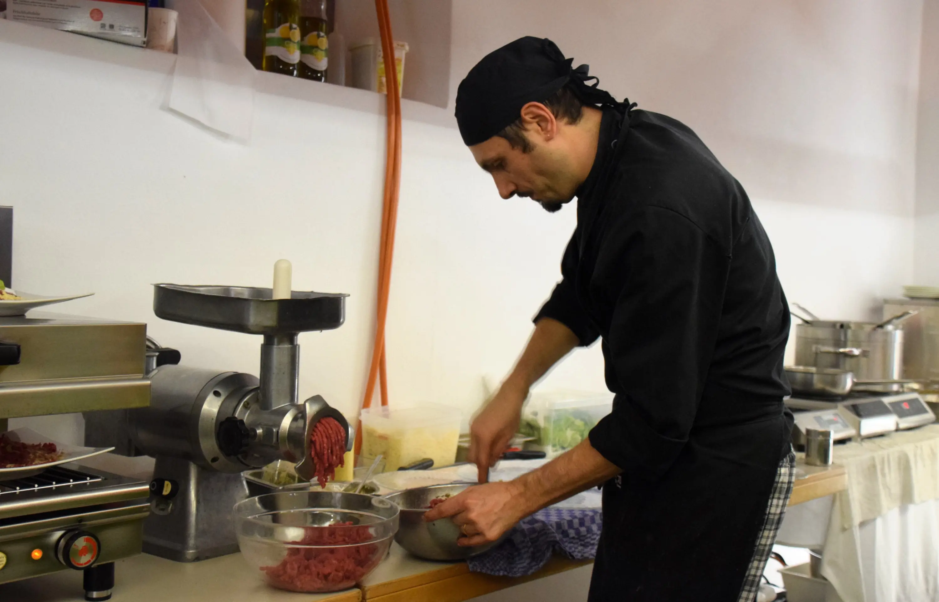 Cuoco al lavoro in una cucina, mentre macina carne con un tritacarne. Indossa un cappello da cuoco e un grembiule nero. Sul tavolo ci sono ciotole e ingredienti vari.