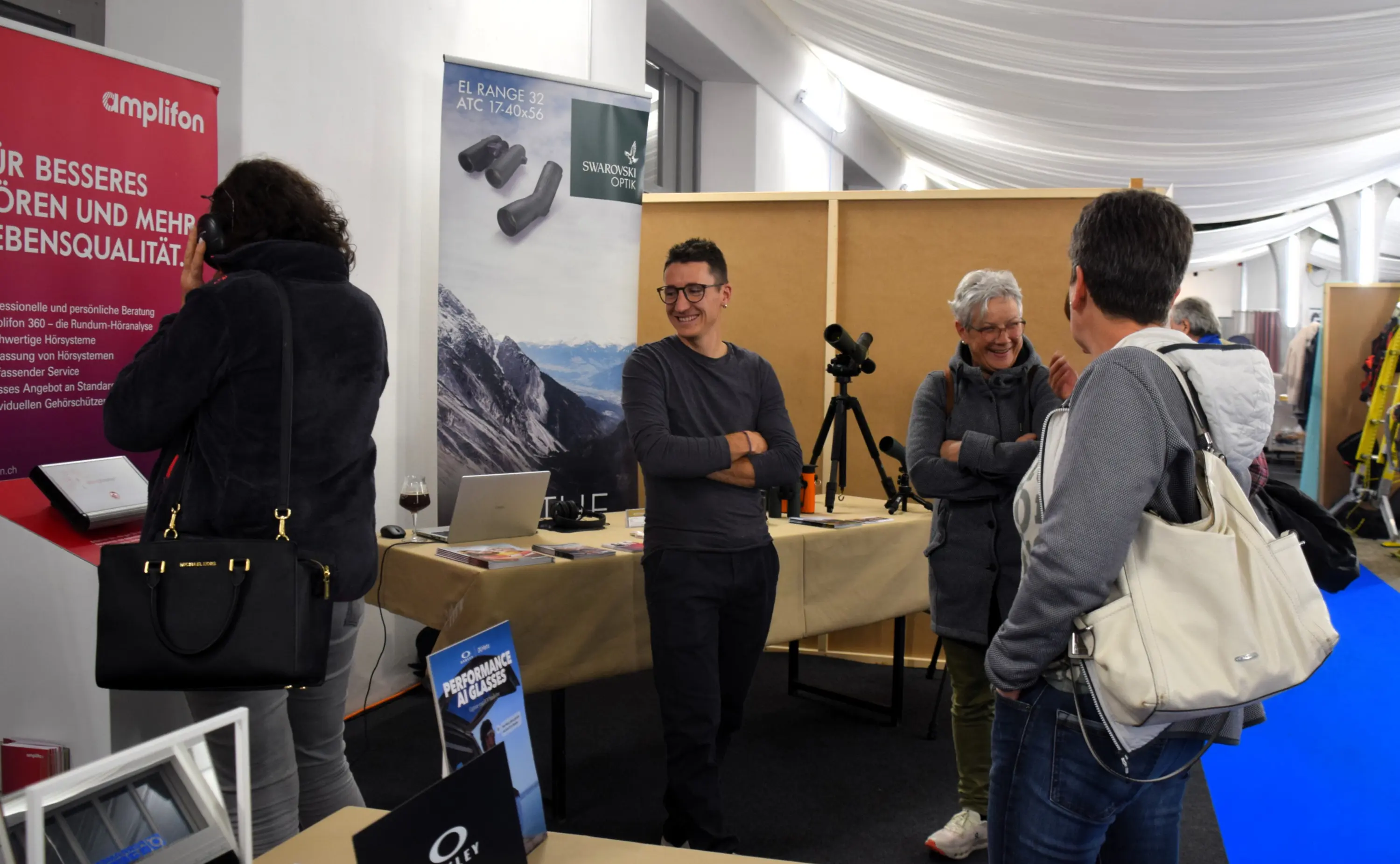 Un gruppo di persone in un evento espositivo, con un tavolo e vari prodotti in mostra. Alcuni partecipanti conversano tra loro e uno sorseggia un bicchiere di vino. In background, materiale promozionale è visibile.
