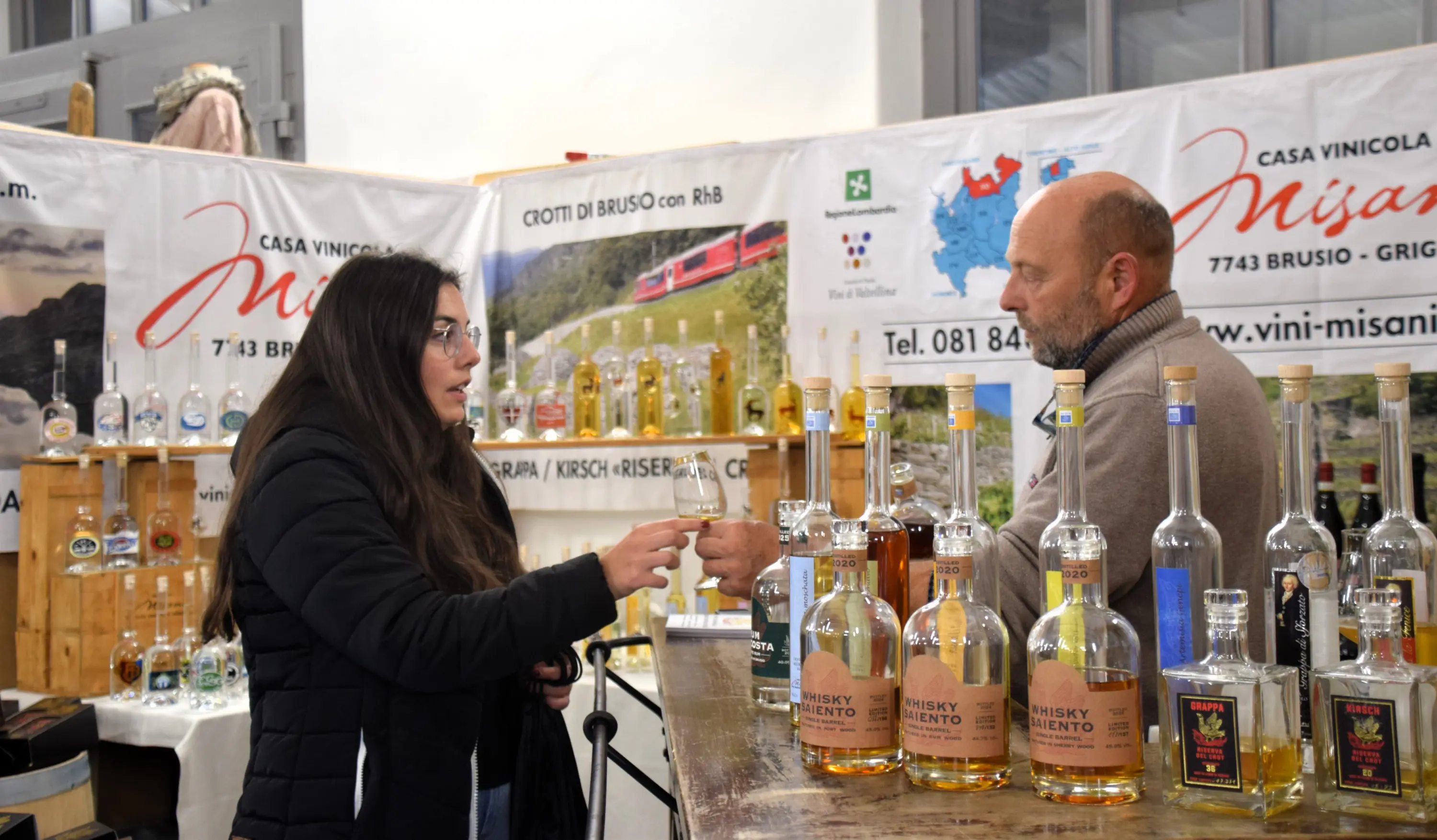 Una donna giovane in giacca nera interagisce con un uomo dietro un bancone, mentre entrambi osservano bottiglie di alcolici. Sullo sfondo ci sono cartelloni pubblicitari e scaffali con varie bottiglie.