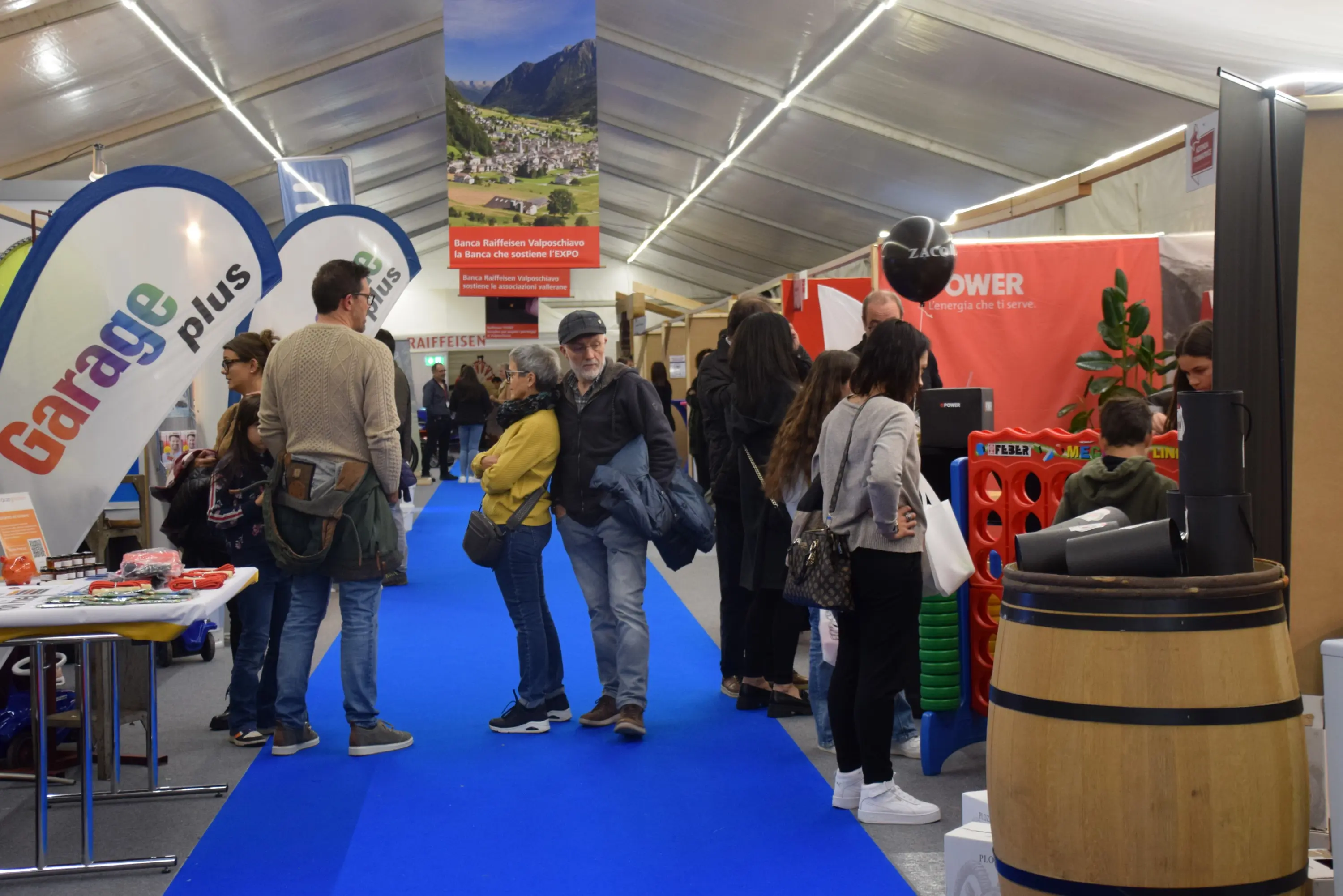 Una fiera con stand espositivi e visitatori in movimento. Alcuni partecipanti osservano prodotti, mentre altri chiacchierano. Si notano decorazioni e banner che pubblicizzano vari servizi. Il pavimento è di un blu acceso.