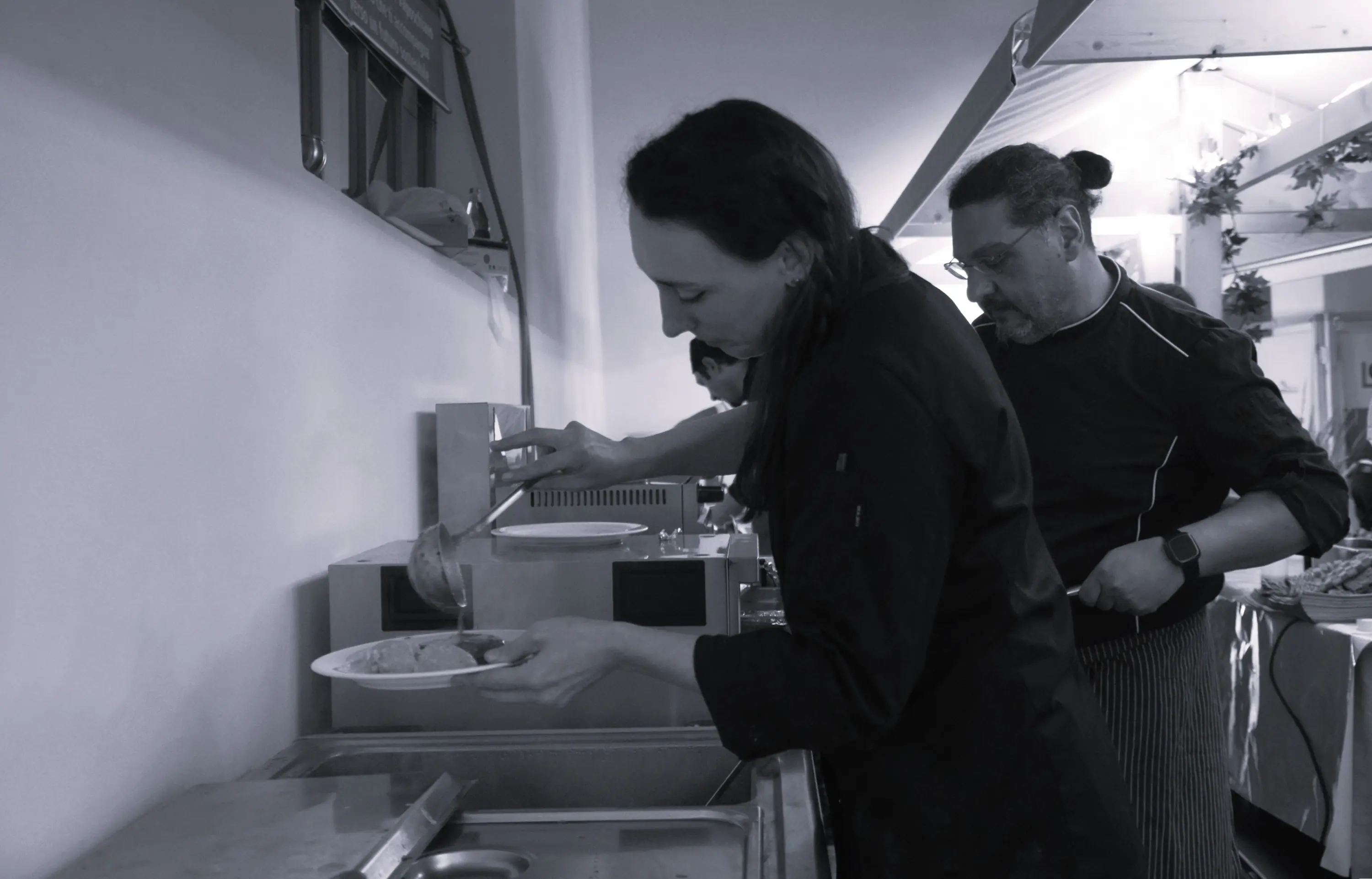 Due chef in cucina mentre servono piatti. Una donna versa il cibo su un piatto, mentre un uomo la osserva con attenzione. L'ambiente è luminoso e ben organizzato.