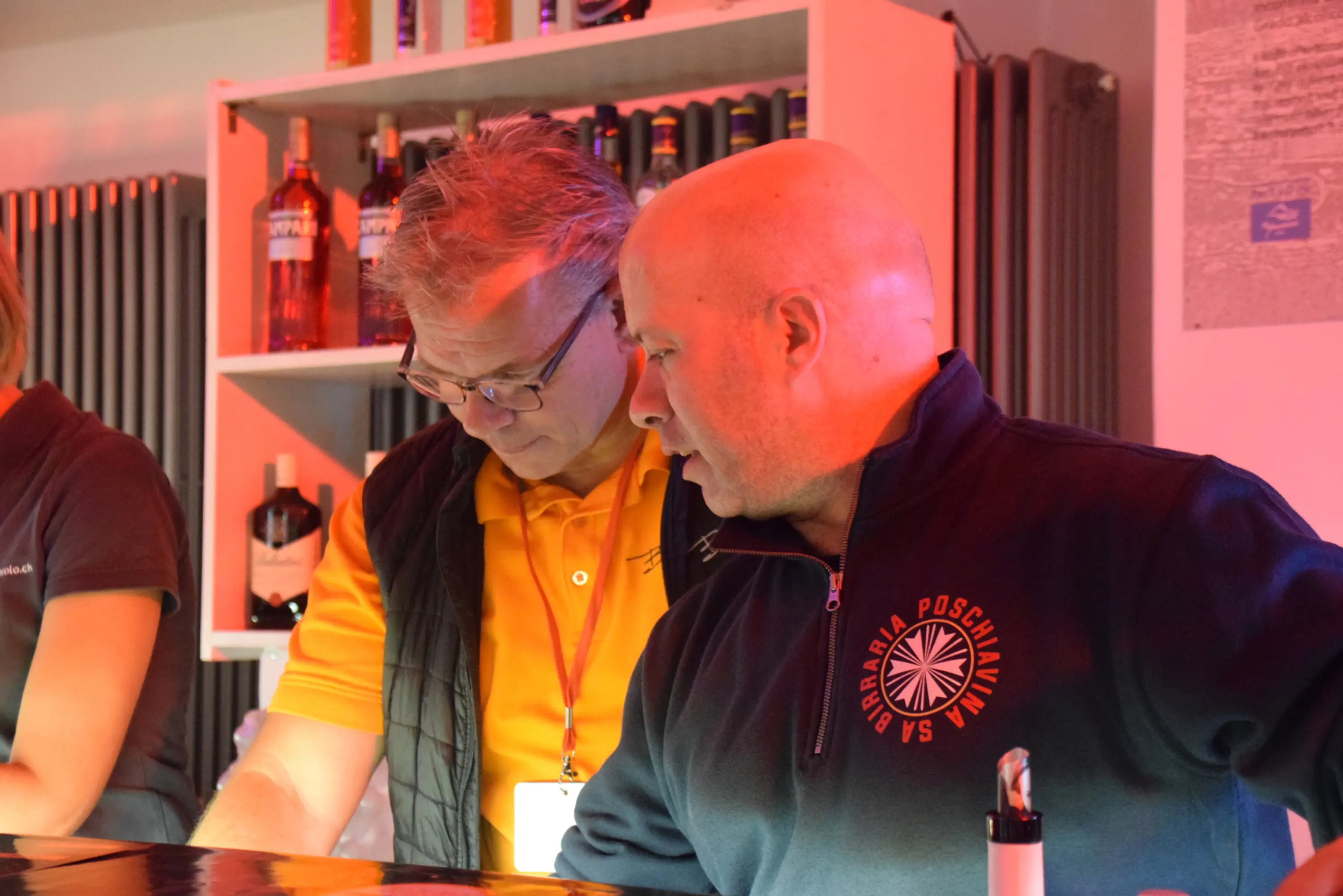 Due uomini al bancone di un bar osservano attentamente un foglio. Uno indossa una maglia nera con una scritta, l'altro una maglietta arancione. Sullo sfondo si vedono bottiglie di liquori illuminate da una luce rossa.