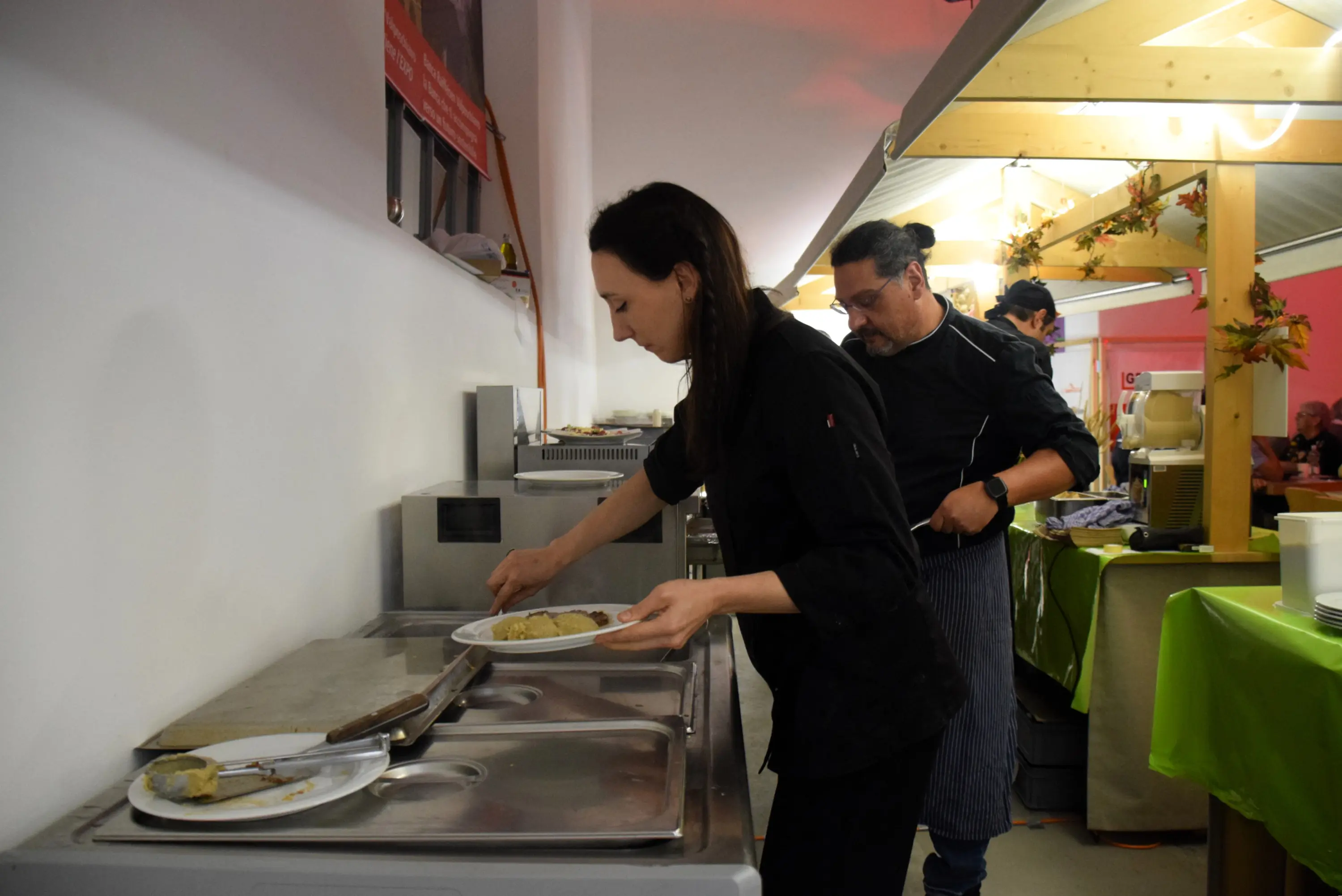 Una donna in abbigliamento da cucina sta impiattando un piatto di cibo in una cucina. Dietro di lei, un uomo con un grembiule osserva mentre cucina. L'ambiente è luminoso, con attrezzature da cucina e decorazioni.