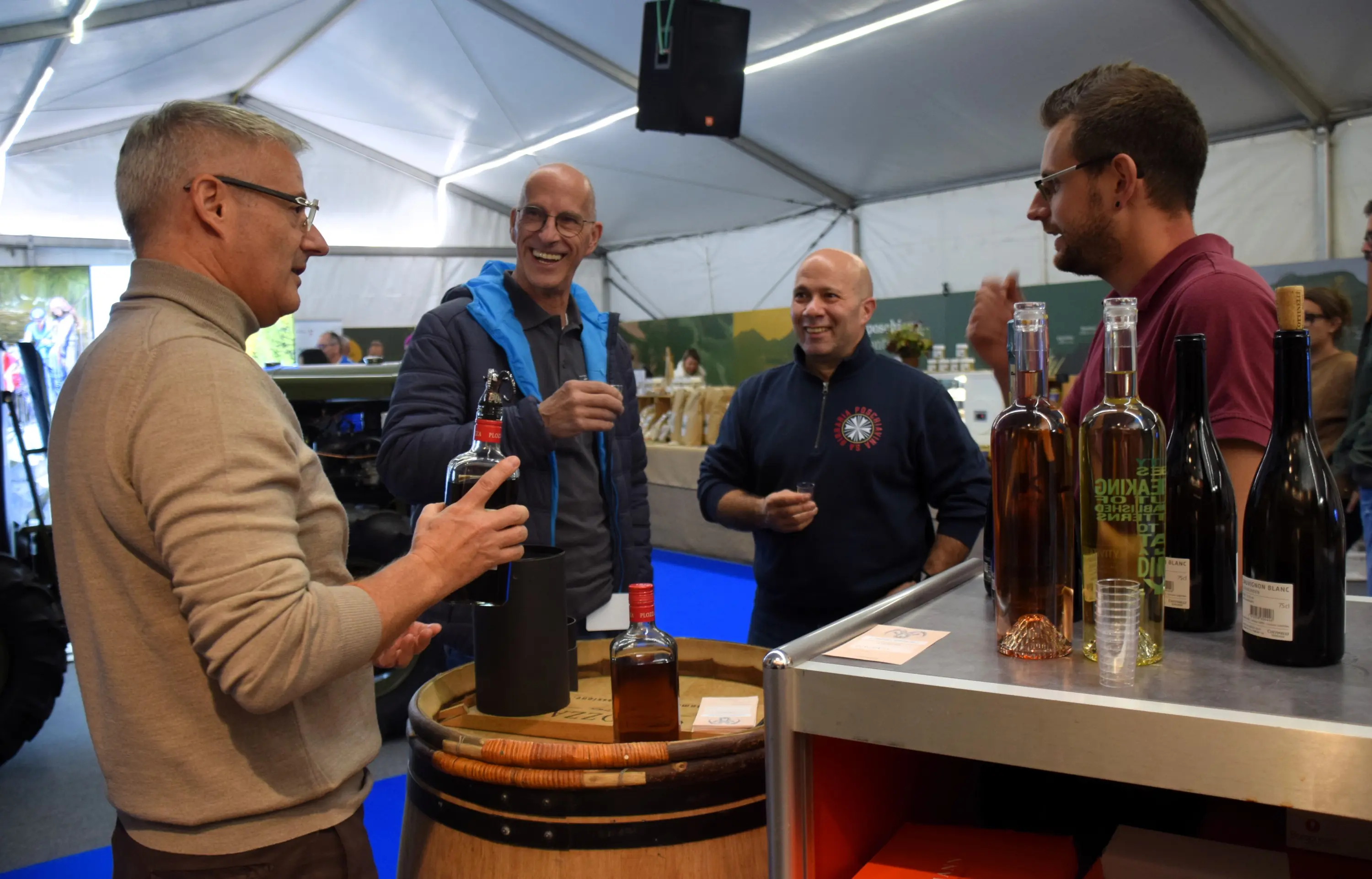 Quattro uomini sorridenti conversano mentre degustano vini all'interno di una tenda per un evento enologico. Uno tiene una bottiglia di vino e un bicchiere, mentre gli altri si scambiano impressioni. Sullo sfondo, bottiglie di vino sono esposte su un tavolo e un grande barile in legno è presente accanto a loro.