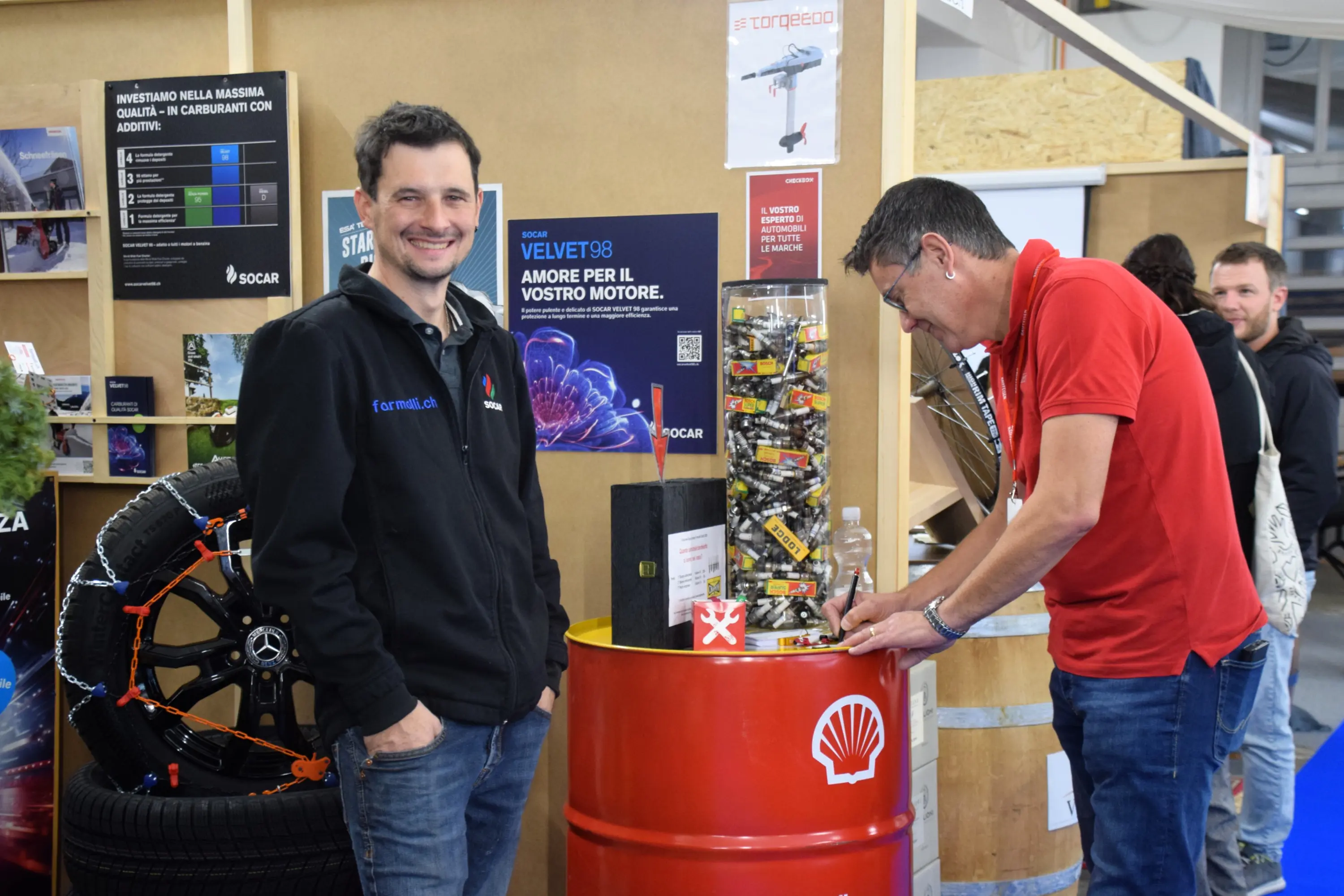 Due uomini in un ambiente fieristico. Uno sorride mentre sta in piedi accanto a un tamburo di Shell, mentre l'altro è chinato e scrive su un modulo. Sullo sfondo ci sono poster e una ruota.