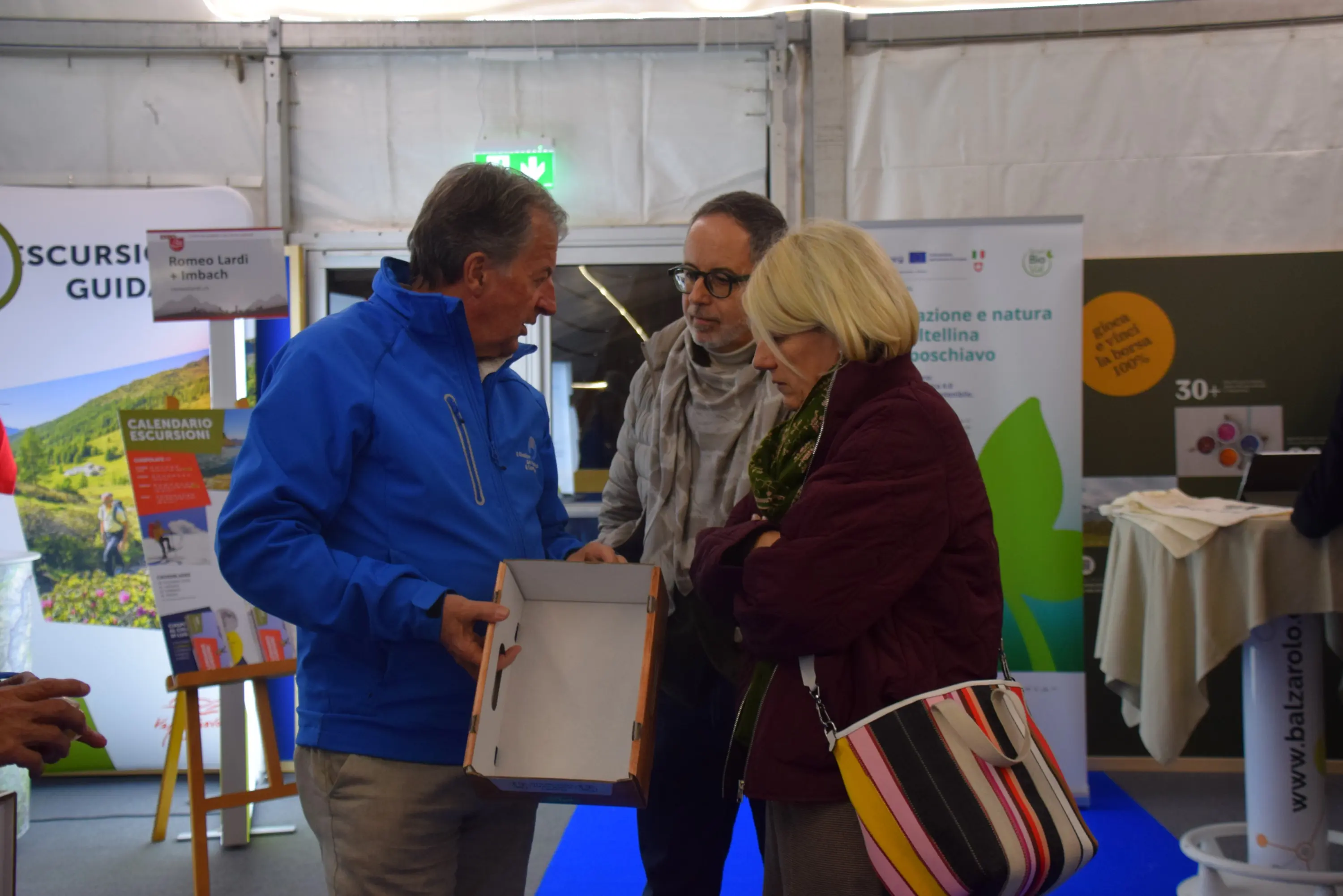 Un uomo in giacca blu mostra una scatola a due persone, una con i capelli chiari e l'altra con occhiali. Sullo sfondo si vedono banner informativi e un'atmosfera di evento.