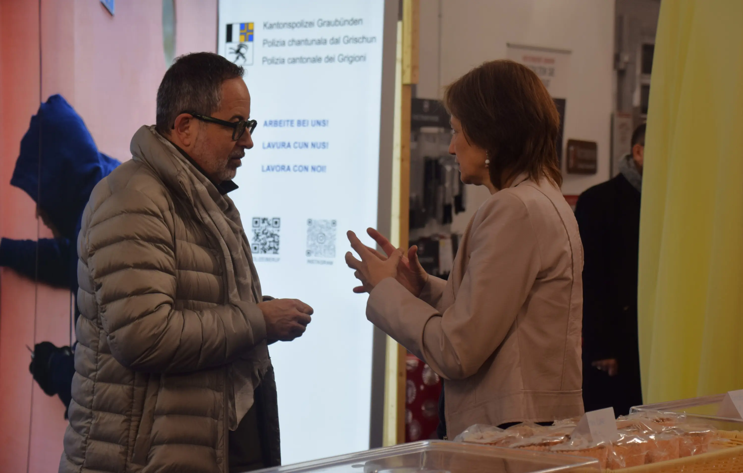 Due persone stanno conversando all'interno di un edificio. Un uomo con occhiali e una giacca argentata ascolta attentamente mentre una donna in giacca beige gesticola. Sullo sfondo si notano cartelli e una vetrina con prodotti.