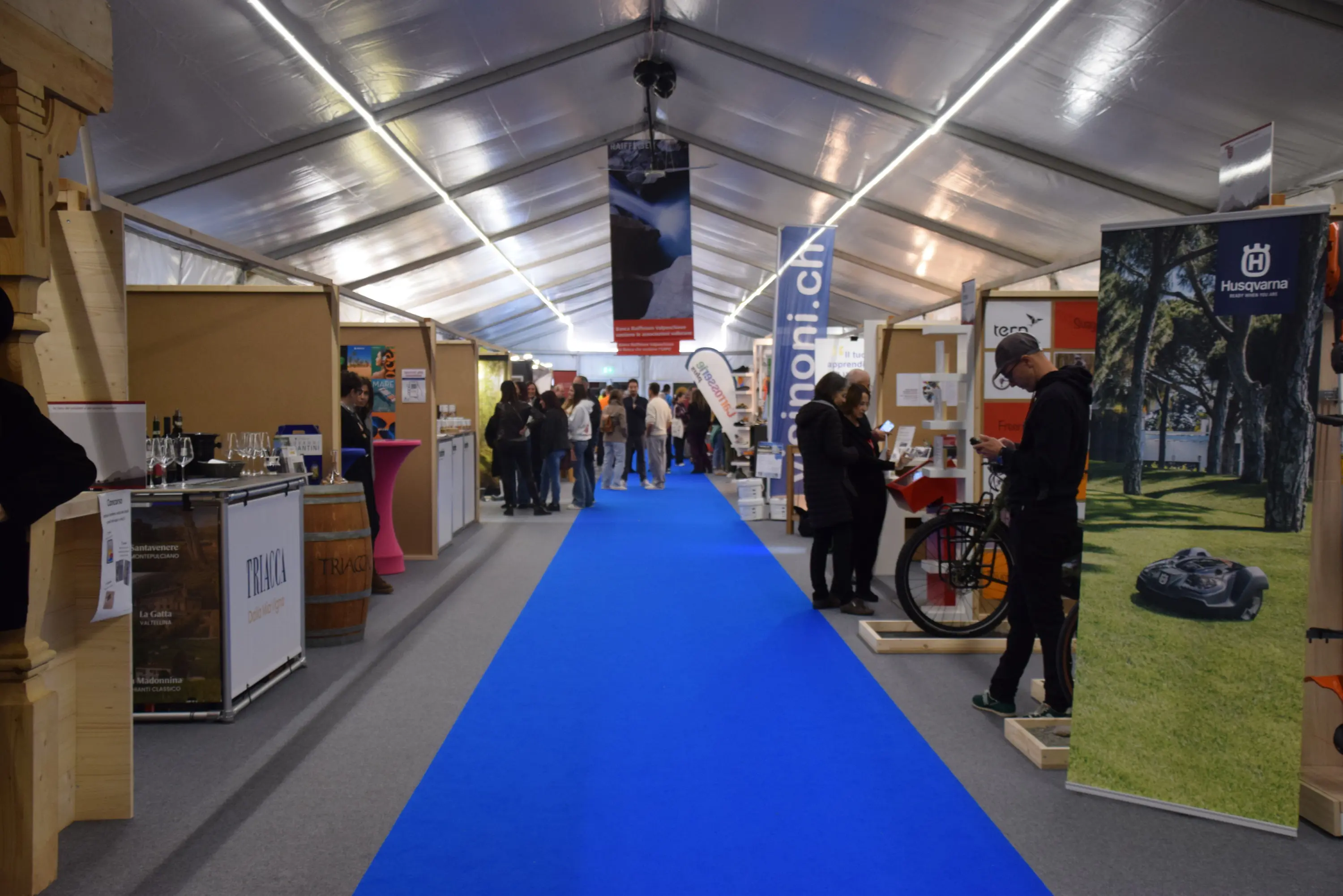 Un'ampia tenda espositiva con un tappeto blu che attraversa il centro. Vari stand mostrano prodotti e servizi, mentre i visitatori si muovono lungo il corridoio. Alcuni espositori mostrano biciclette e tecnologia per il giardinaggio.
