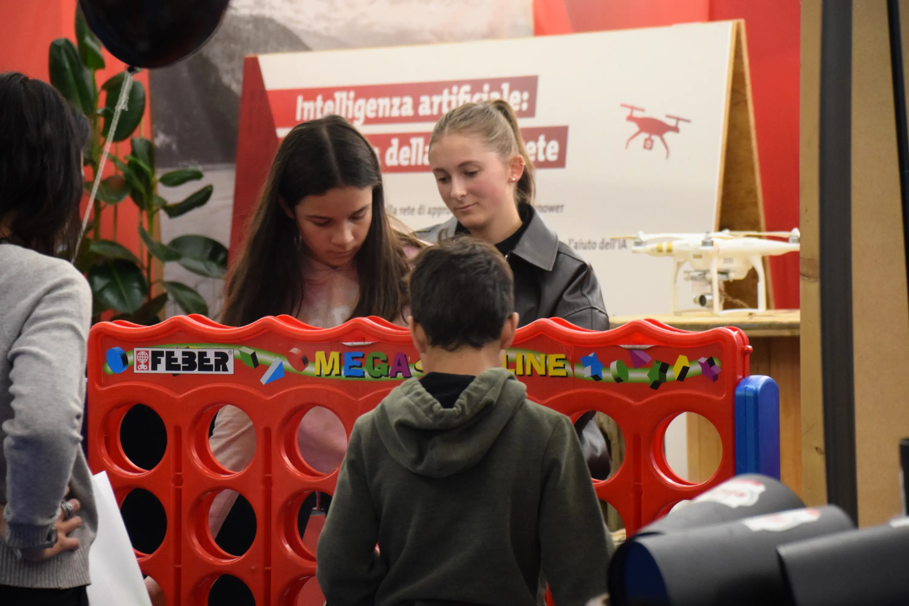 Due ragazze interagiscono con un gioco da tavolo di collegamento, mentre un bambino osserva. Sullo sfondo si trova un drone e un cartello sull'intelligenza artificiale.