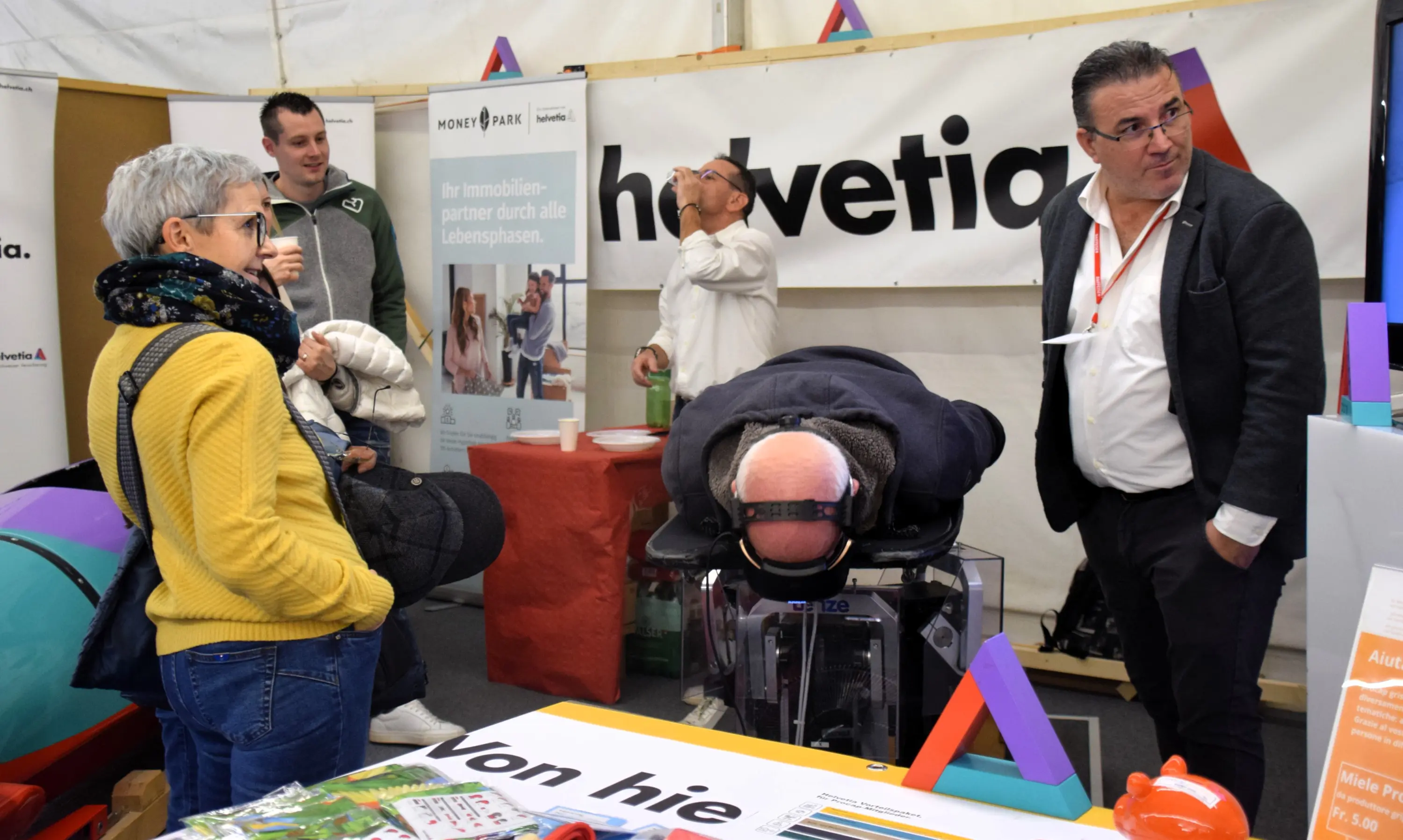 Un gruppo di persone in un evento, alcune osservano un uomo sdraiato su un dispositivo di salute. Sono presenti poster e materiale promozionale sullo sfondo.