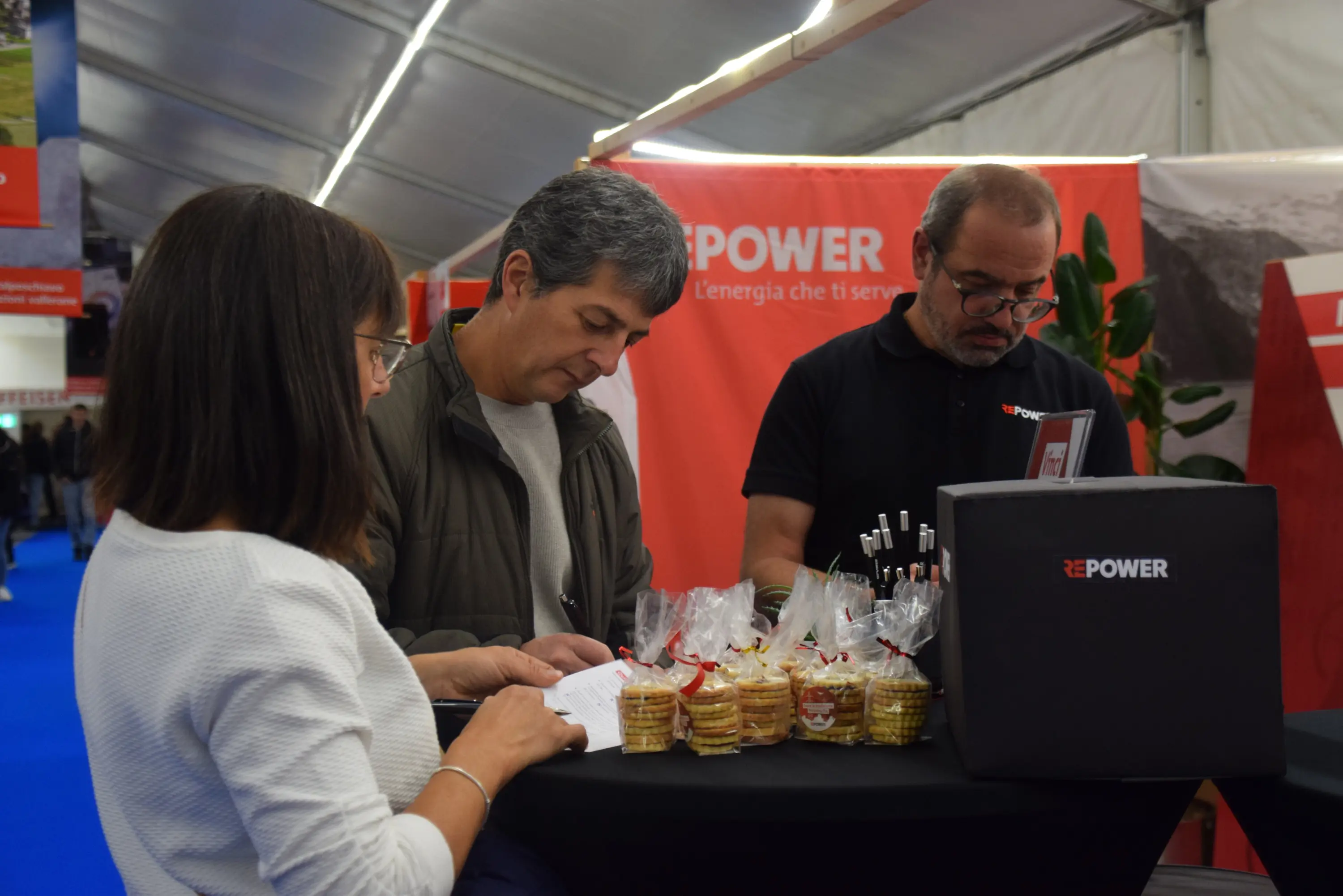 Tre persone si trovano a un tavolo in un evento, mentre interagiscono tra di loro. Due sono in primo piano: una donna che scrive e un uomo che legge qualcosa. L'uomo sullo sfondo tiene una penna e sorride. Sul tavolo ci sono sacchetti con dolcetti e un pacco nero di REPOWER.