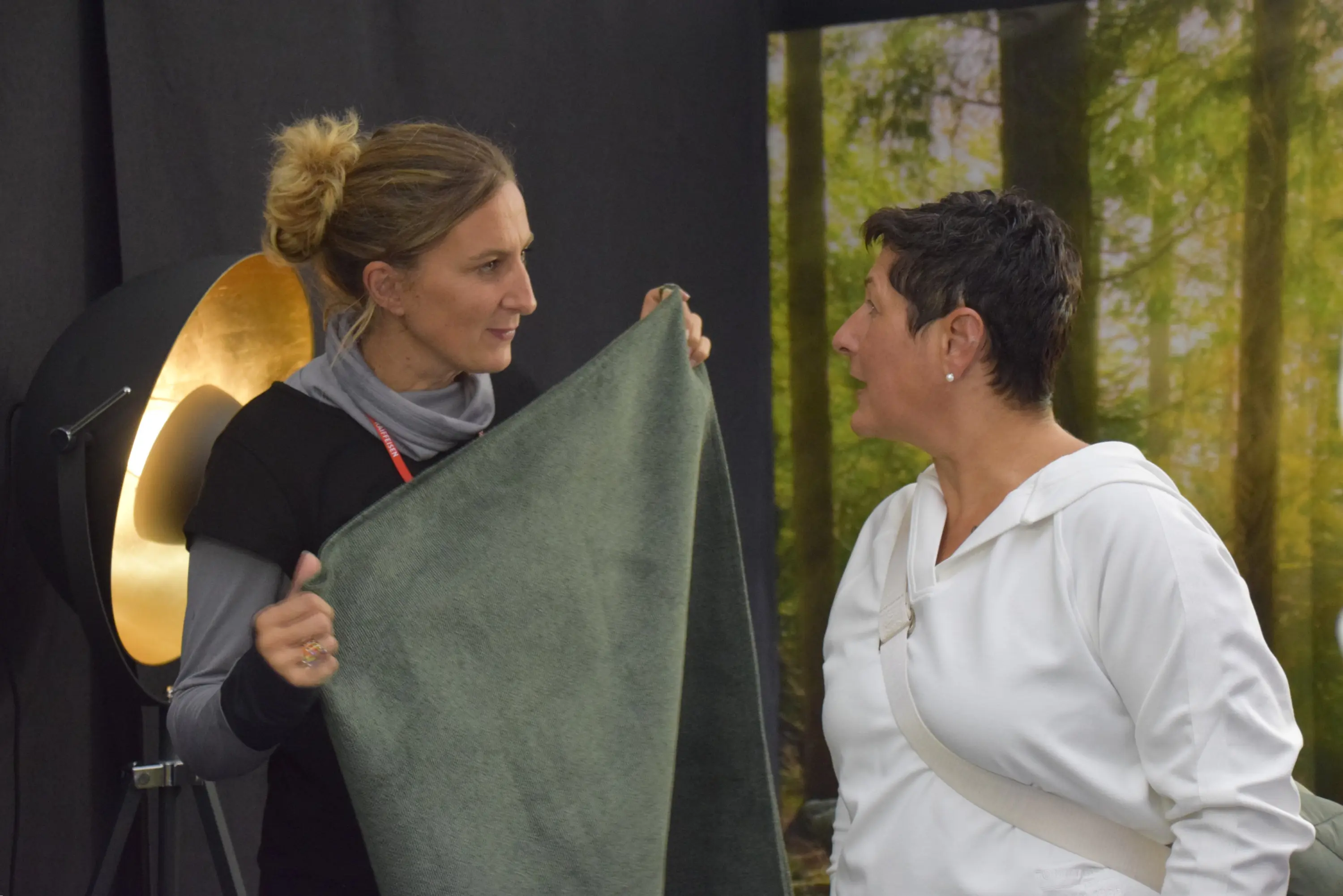 Due donne stanno conversando, una tiene un tessuto verde mentre l'altra ascolta attentamente. Sullo sfondo si intravede un'illuminazione soffusa e un'immagine di un bosco.