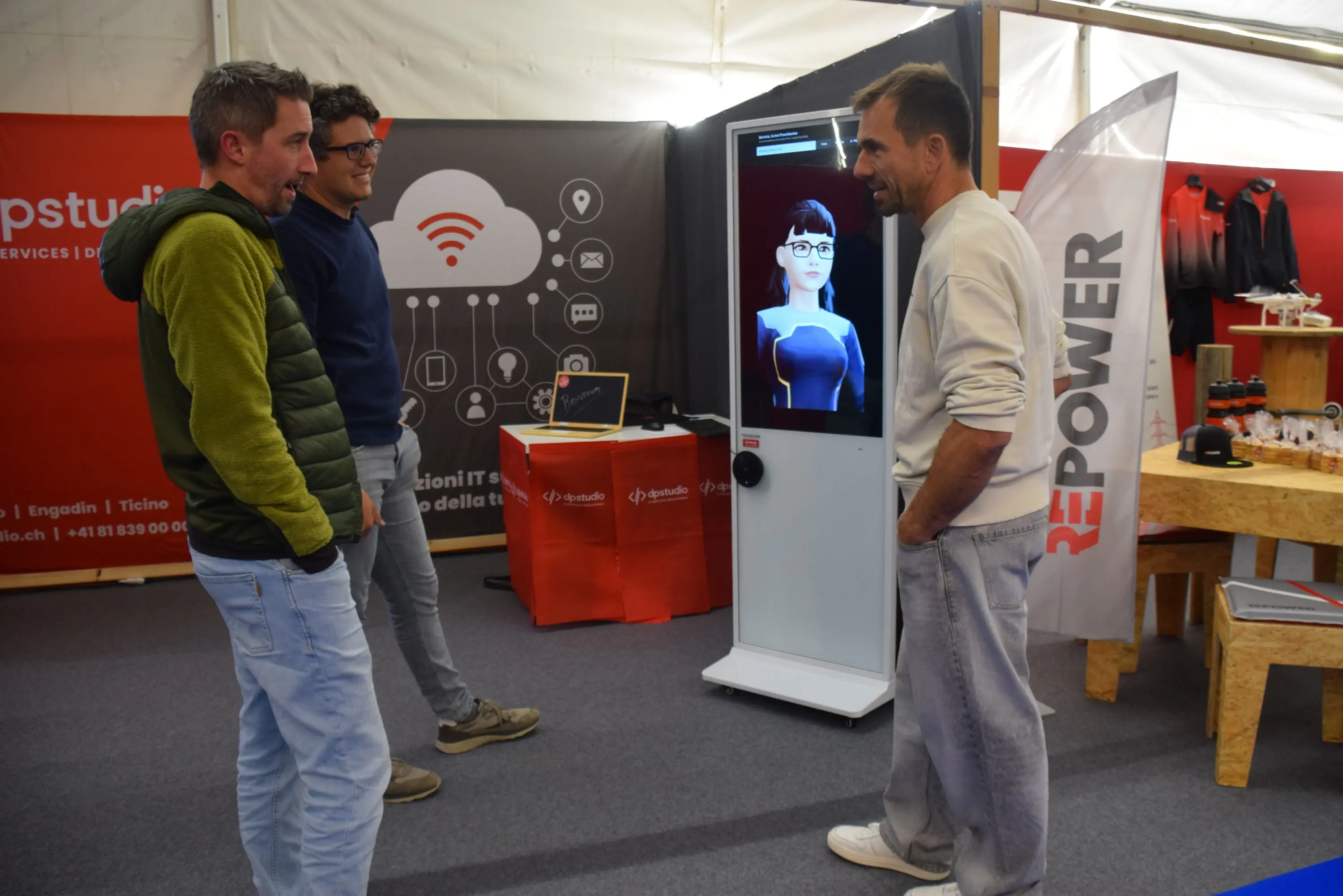 Due uomini conversano accanto a un chiosco interattivo con un avatar virtuale. Un altro uomo osserva la scena. Lo sfondo presenta banner e stand commerciali.