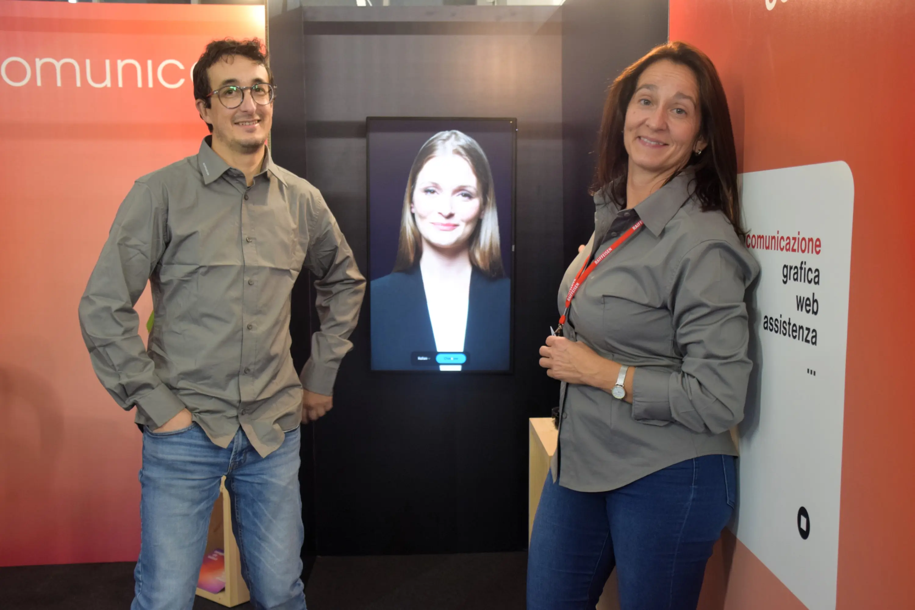 Due persone sorridenti in abiti casual si trovano davanti a un pannello pubblicitario. Dietro di loro, uno schermo mostra il volto di una donna. I due sembrano rappresentare un'azienda che offre servizi di comunicazione.