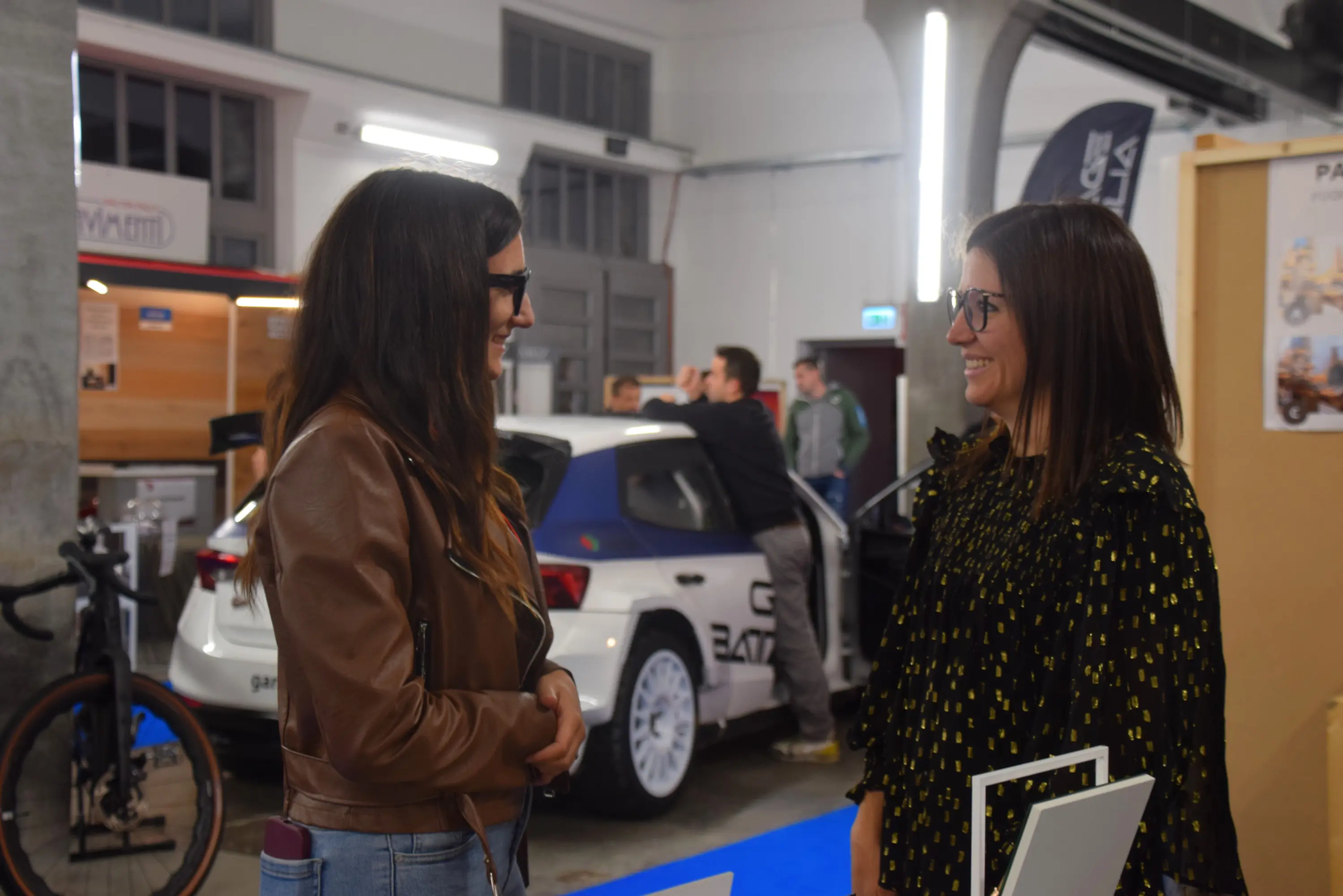 Due donne sorridenti chiacchierano in un ambiente fieristico, con un'auto sportiva sullo sfondo. In primo piano, una donna con occhiali e giacca di pelle parla con un'altra donna in abbigliamento nero decorato. Altre persone si muovono sullo sfondo.