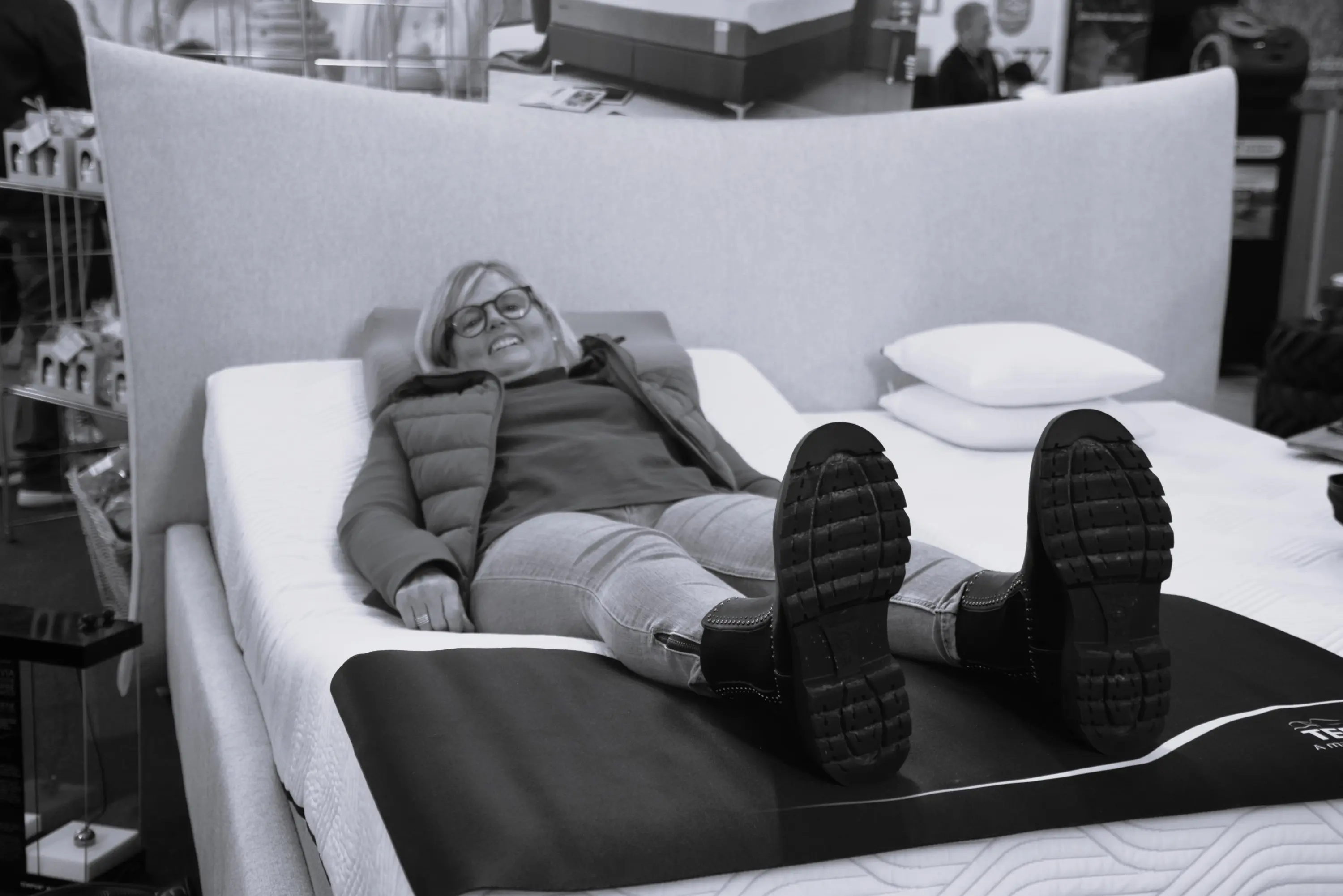 Una donna sorride sdraiata su un materasso in un negozio, con gambe distese e stivali neri.