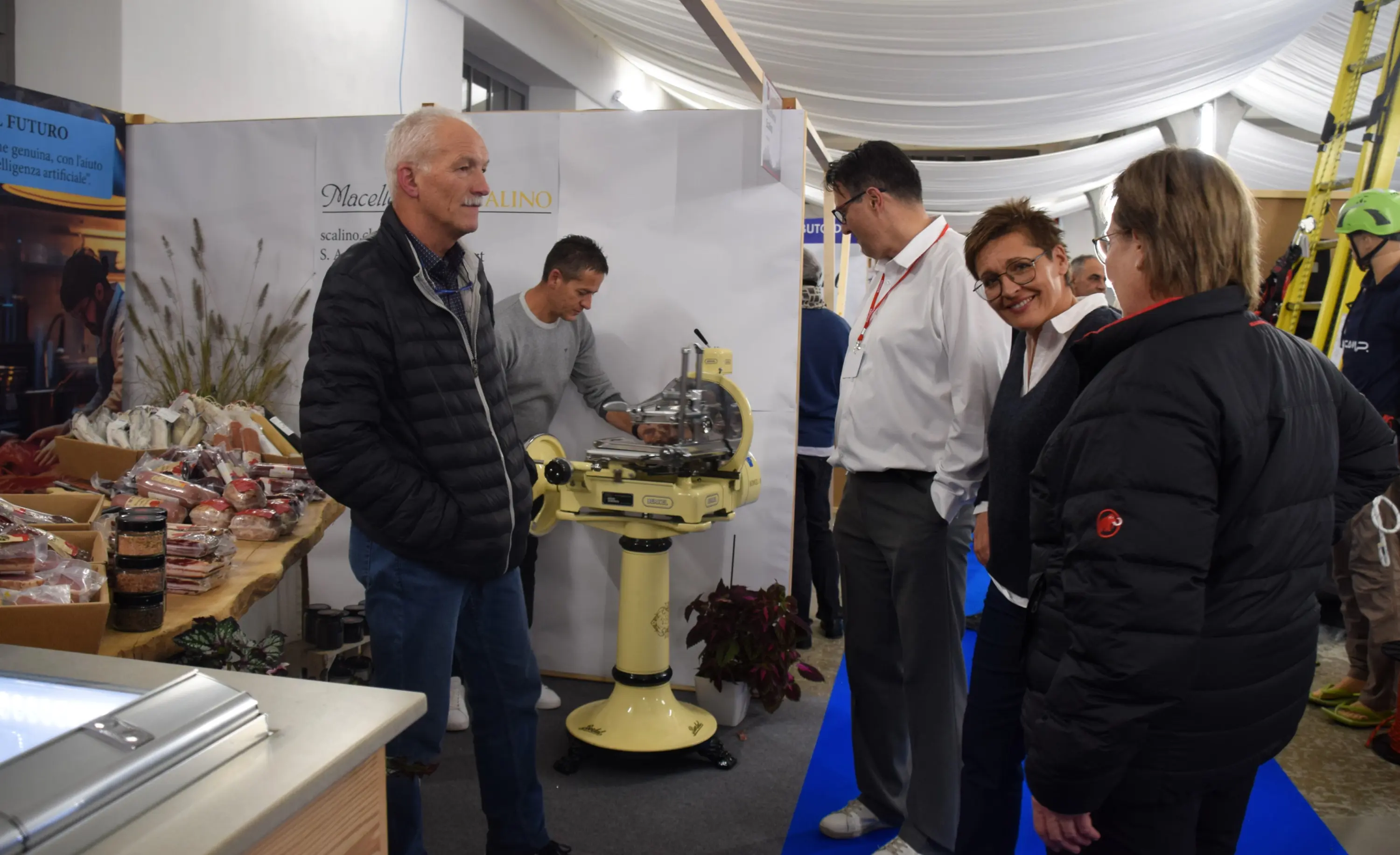Un gruppo di persone interagisce in un evento espositivo. Un uomo con giacca nera parla con alcuni visitatori vicino a una macchina per la lavorazione degli alimenti, mentre altri esplorano un bancone con prodotti alimentari.