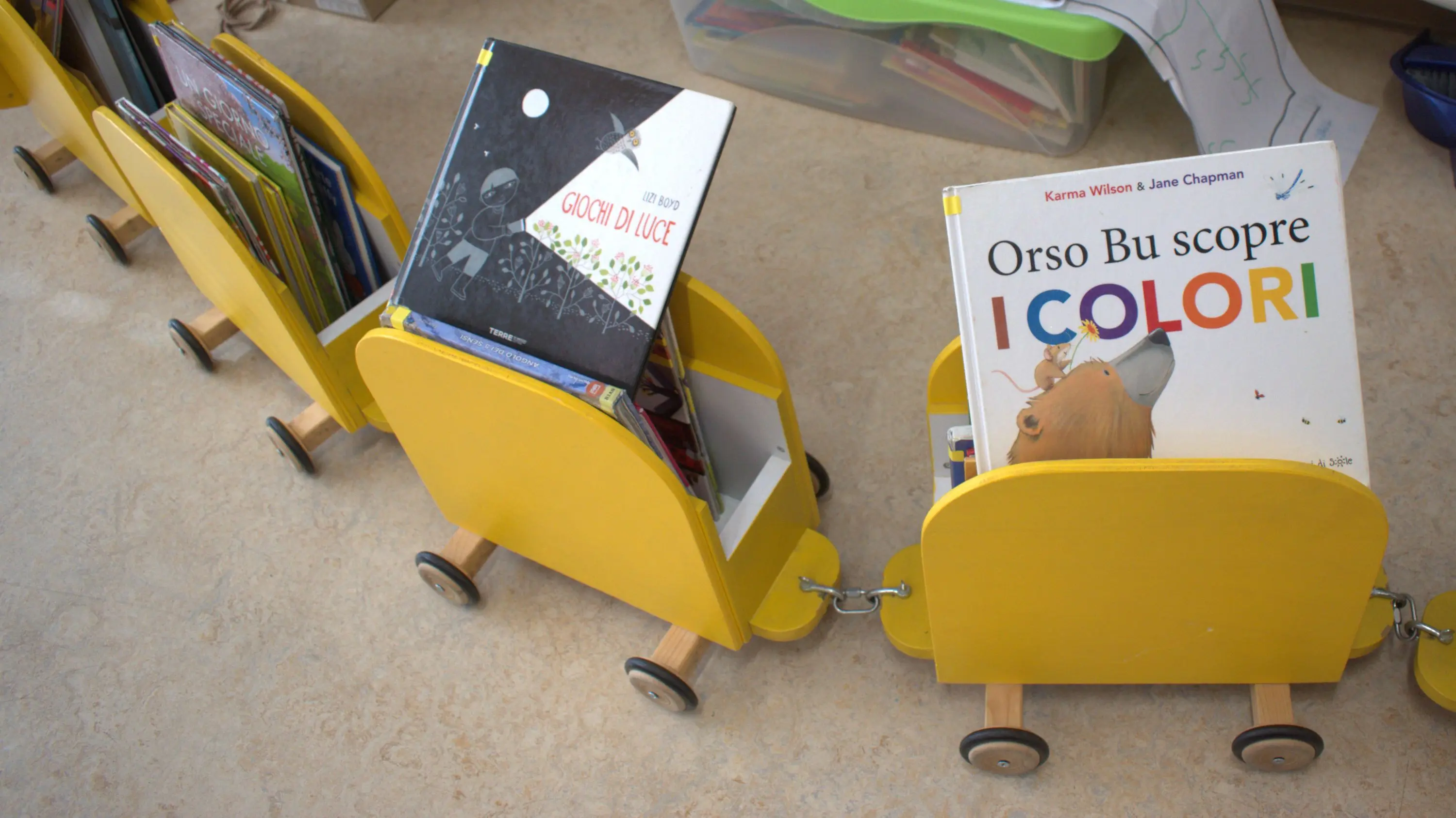 Carrelli gialli con libri per bambini, posizionati su ruote. Due titoli visibili: "Orso Bu scopre I colori" e "Gocce di Luce".