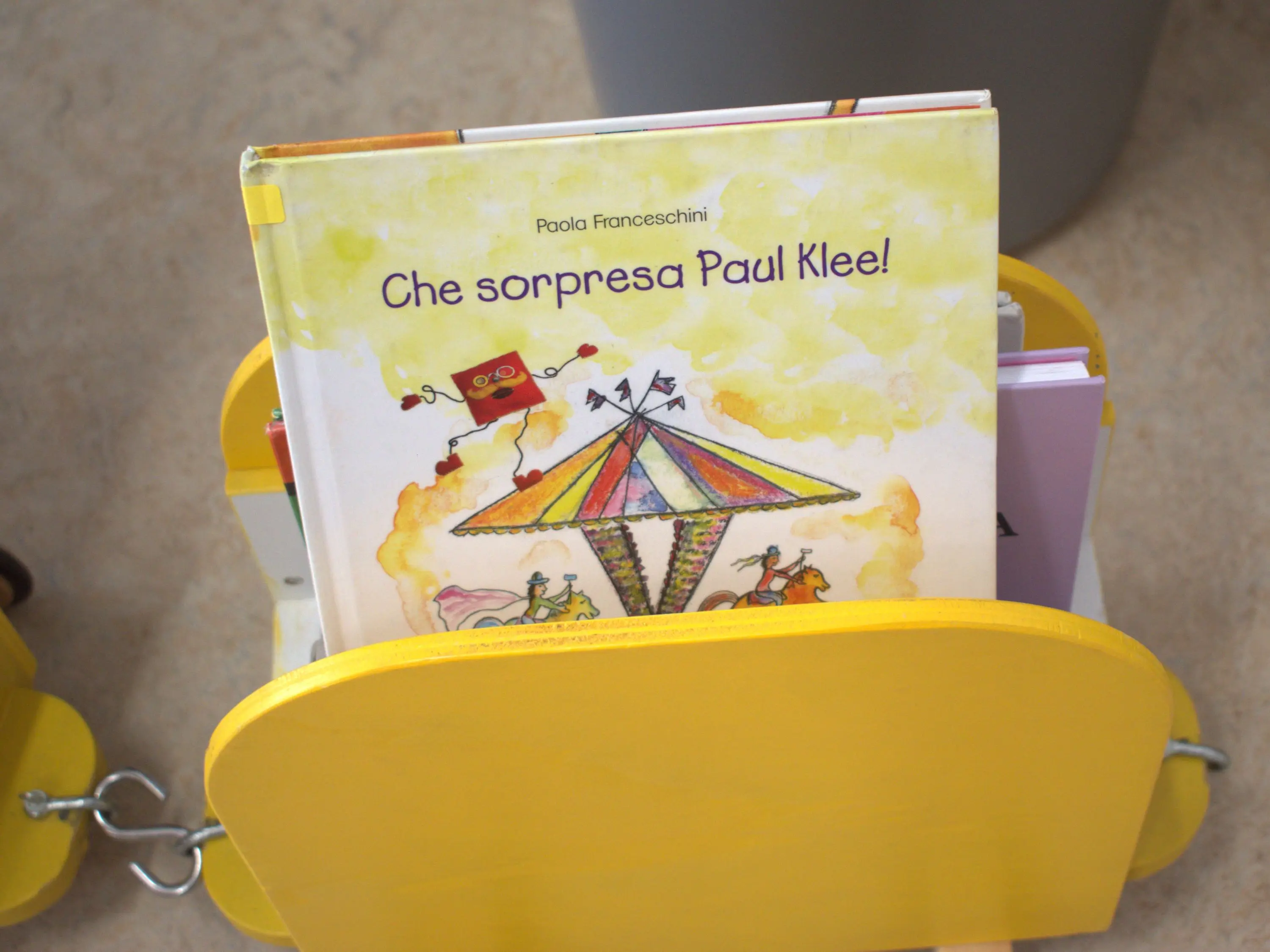 Un libro intitolato "Che sorpresa Paul Klee!" di Paola Franceschini, riposto in un supporto giallo con illustrazioni colorate di un circo e personaggi divertenti.