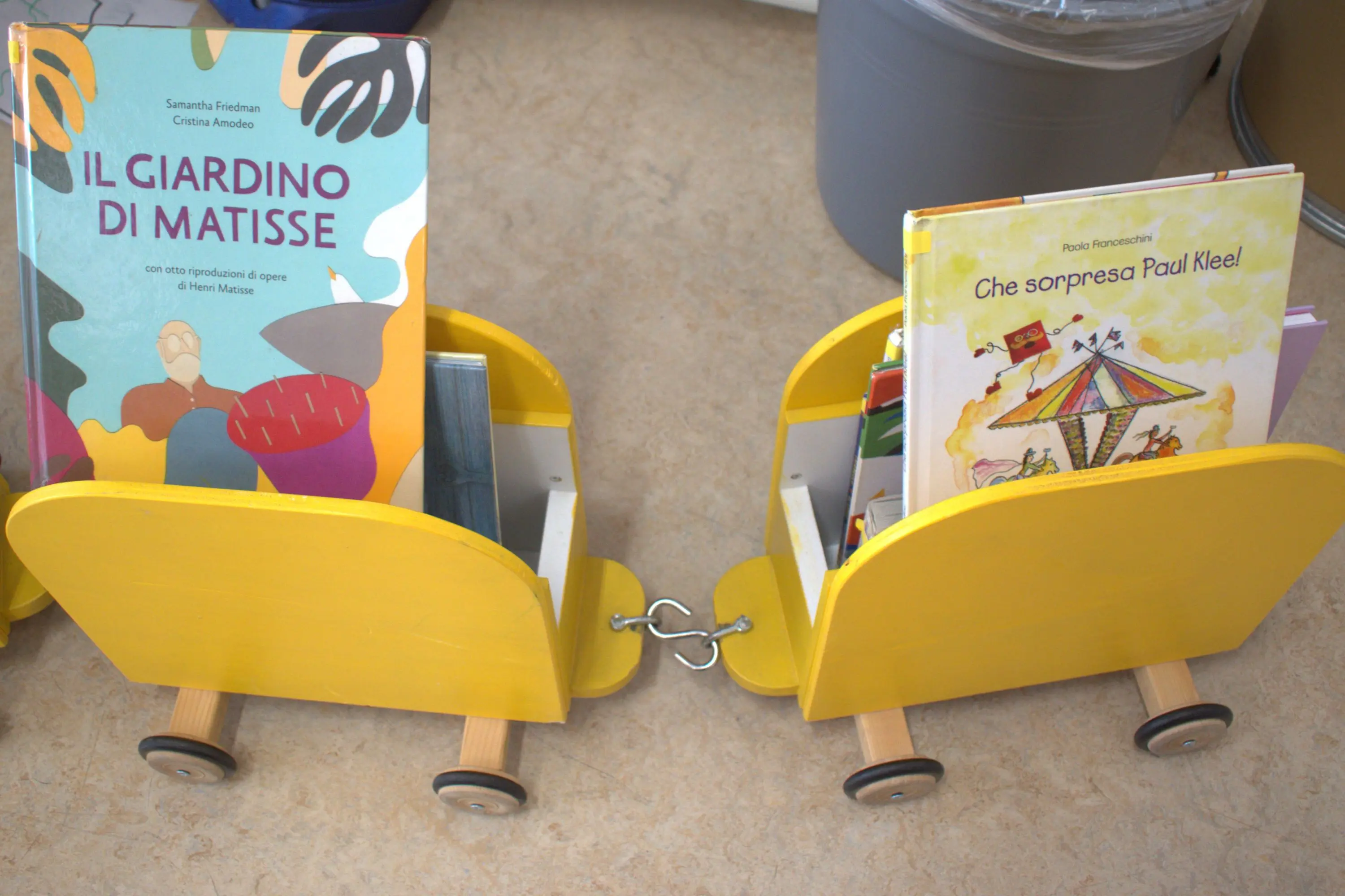 Due carrelli gialli per libri, ciascuno con diverse copertine di libri. A sinistra si trova "Il giardino di Matisse" e a destra "Che sorpresa Paul Klee".