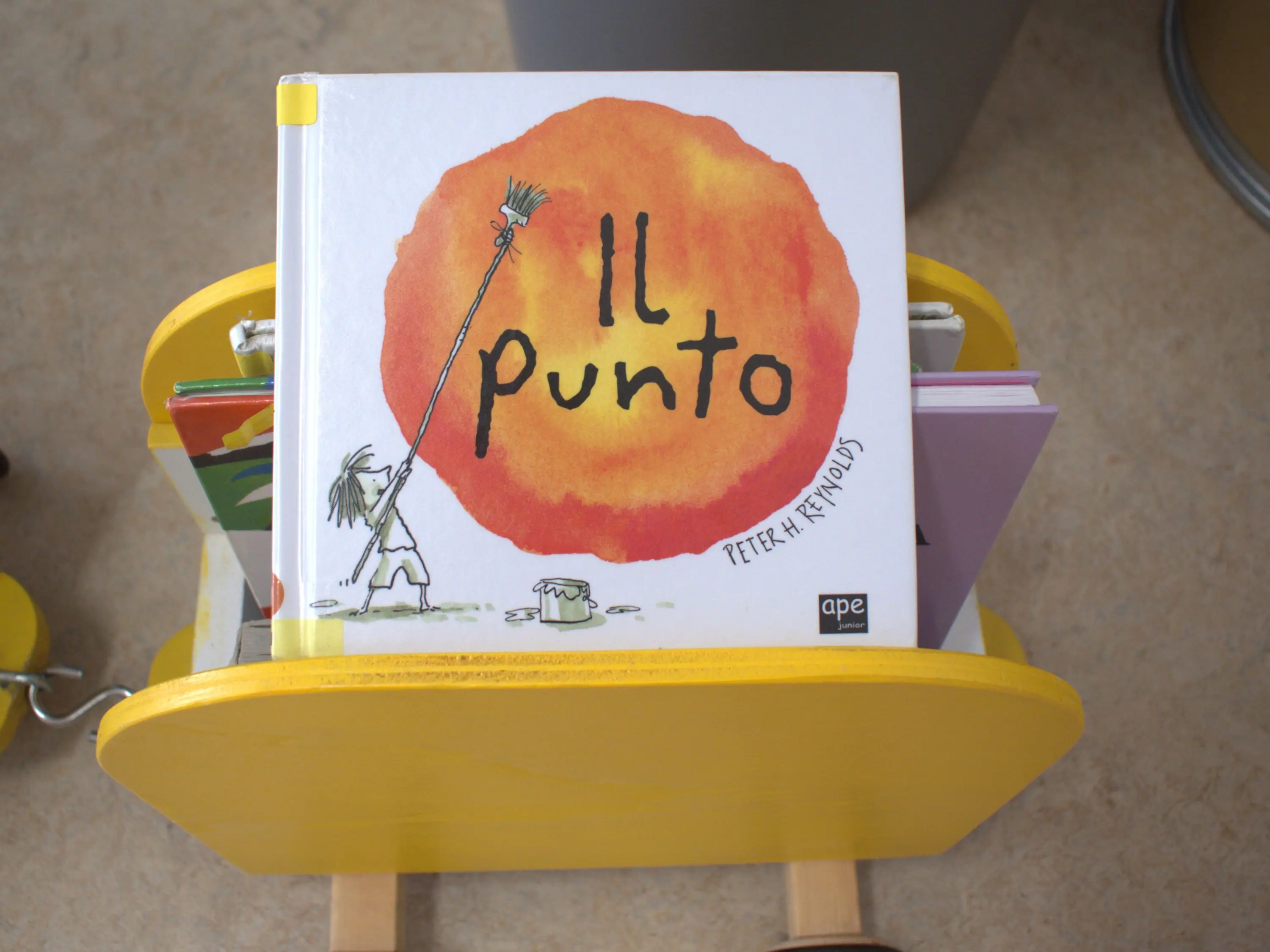 Un libro intitolato "Il punto" di Peter H. Reynolds, con un'illustrazione di un ragazzo che tiene un pennello, di fronte a un grande cerchio arancione. Il libro è posizionato su uno scaffale giallo.