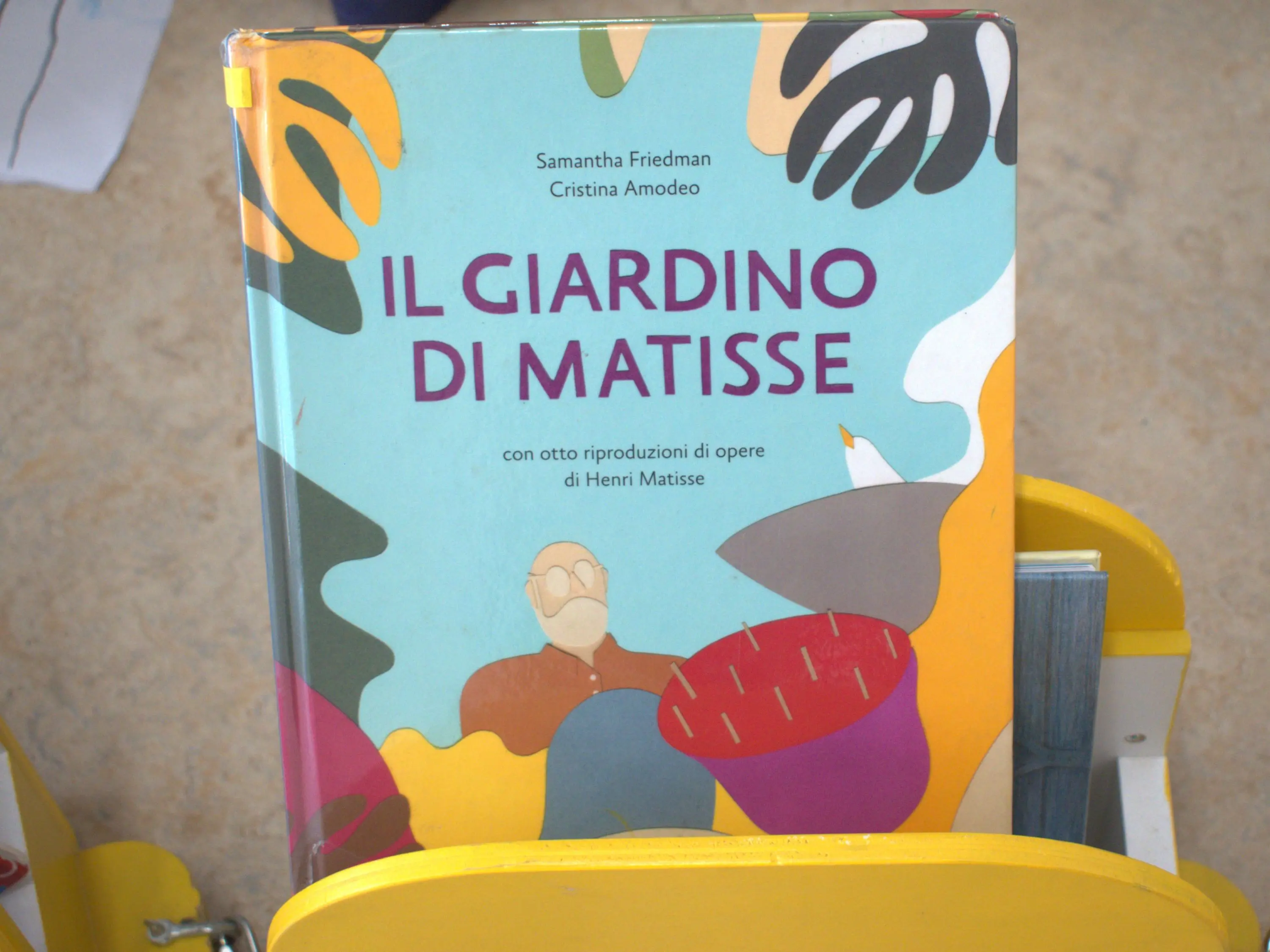 Un libro intitolato "Il Giardino di Matisse", con illustrazioni colorate sulla copertina, è appoggiato su un supporto giallo.