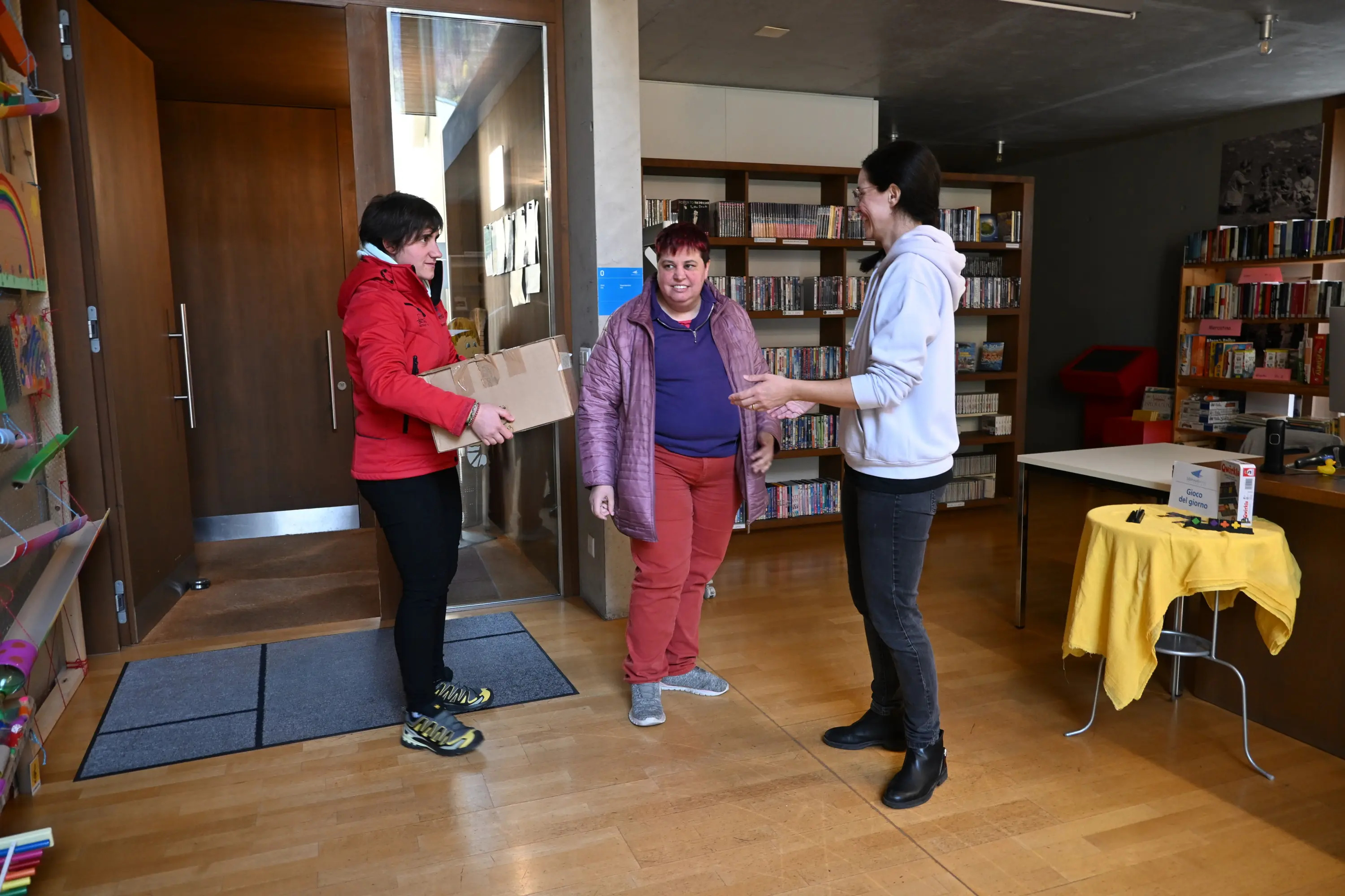 Due donne, una con una giacca rossa e l'altra con una giacca lilla, si trovano all'ingresso di una biblioteca. La donna con la giacca rossa tiene un pac
