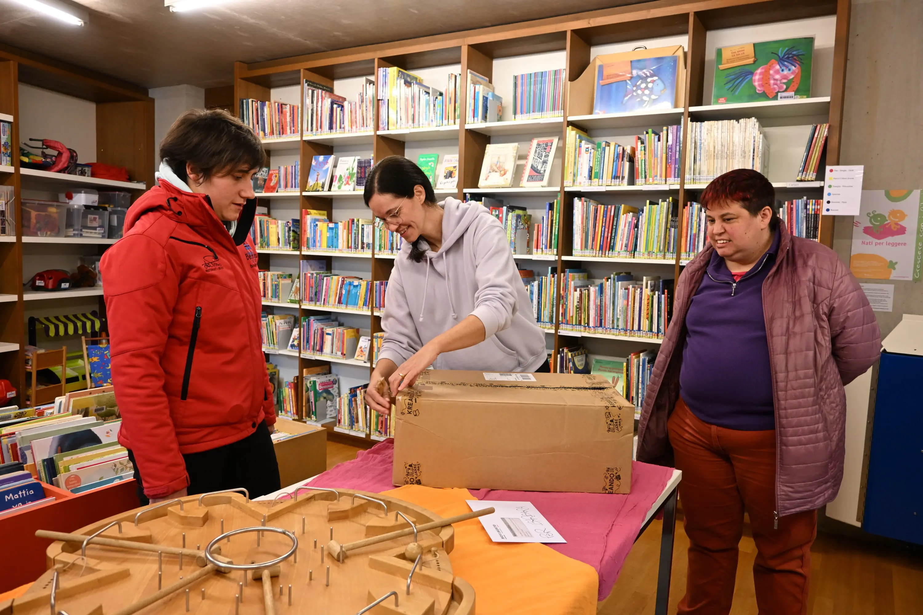 Tre persone stanno lavorando insieme in una biblioteca. Una donna sta aprendo una scatola su un tavolo, mentre le altre due la osservano. Gli scaffali dietro di loro sono pieni