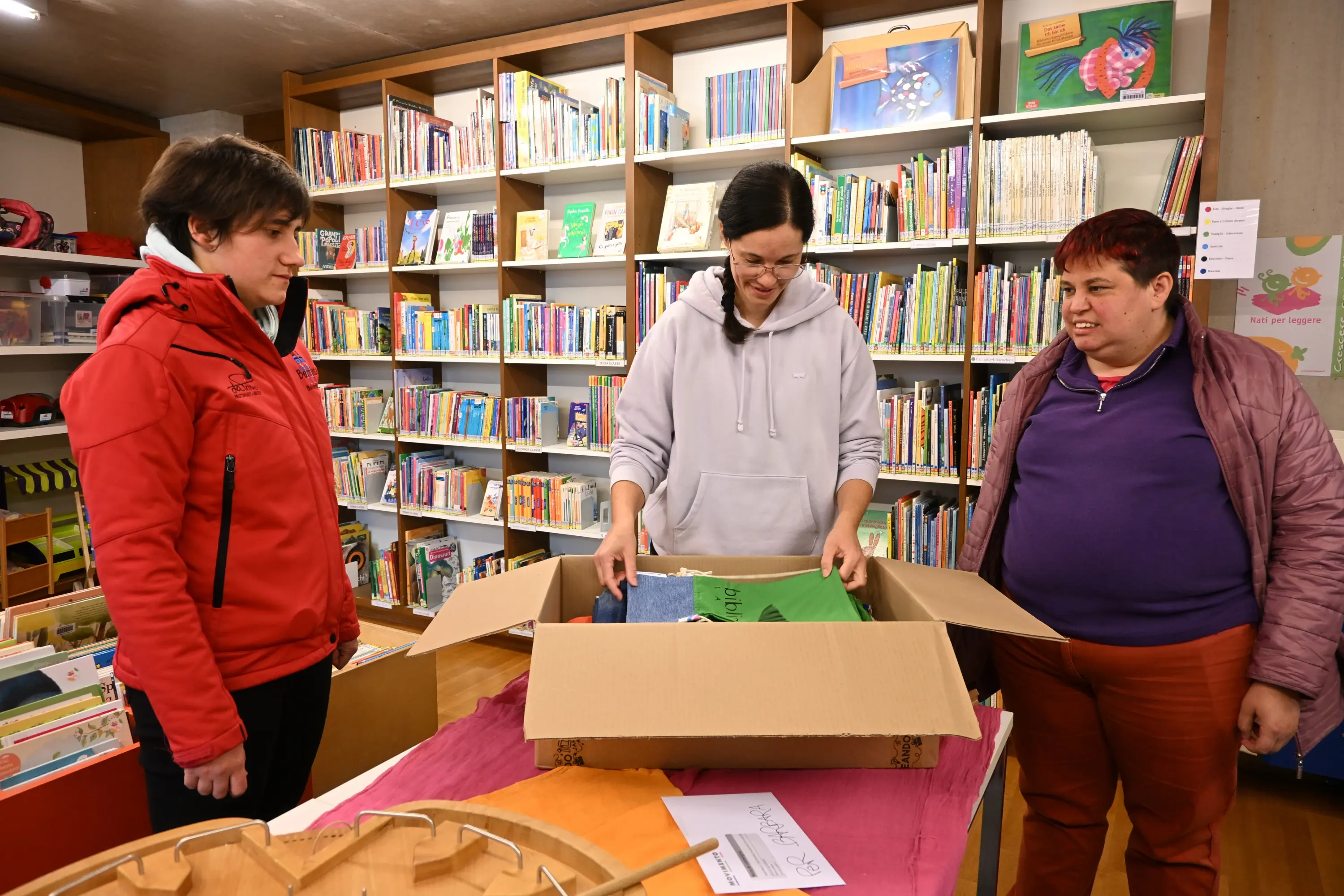 Tre adulti in una biblioteca stanno aprendo una scatola di cartone. Sullo sfondo, ci sono scaffali pieni di libri e materiali didattici. Due delle donne indossano