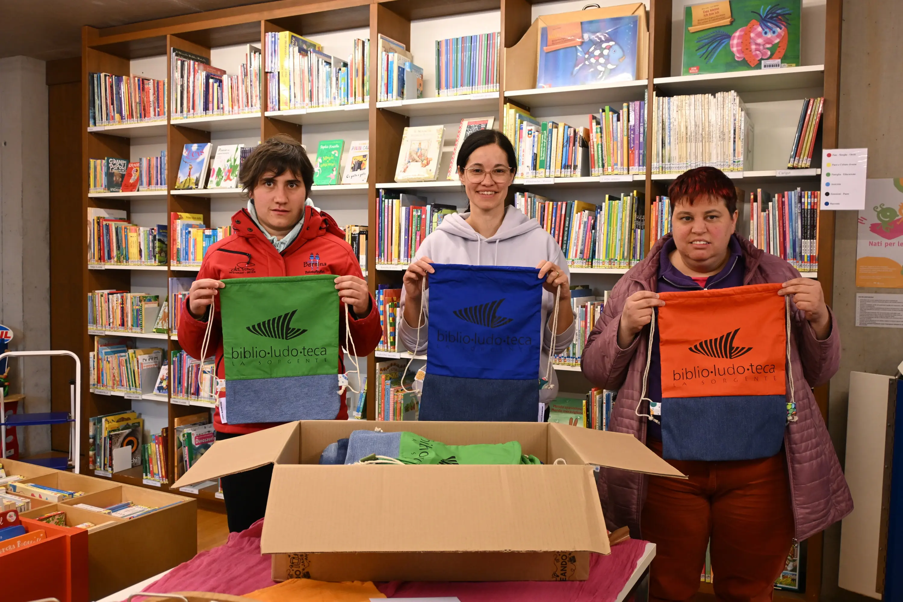 Tre persone sorridenti mostrano borse colorate di diverse tinte in una biblioteca. Dietro di loro si vede uno scaffale pieno di libri. Una di loro tiene una borsa blu