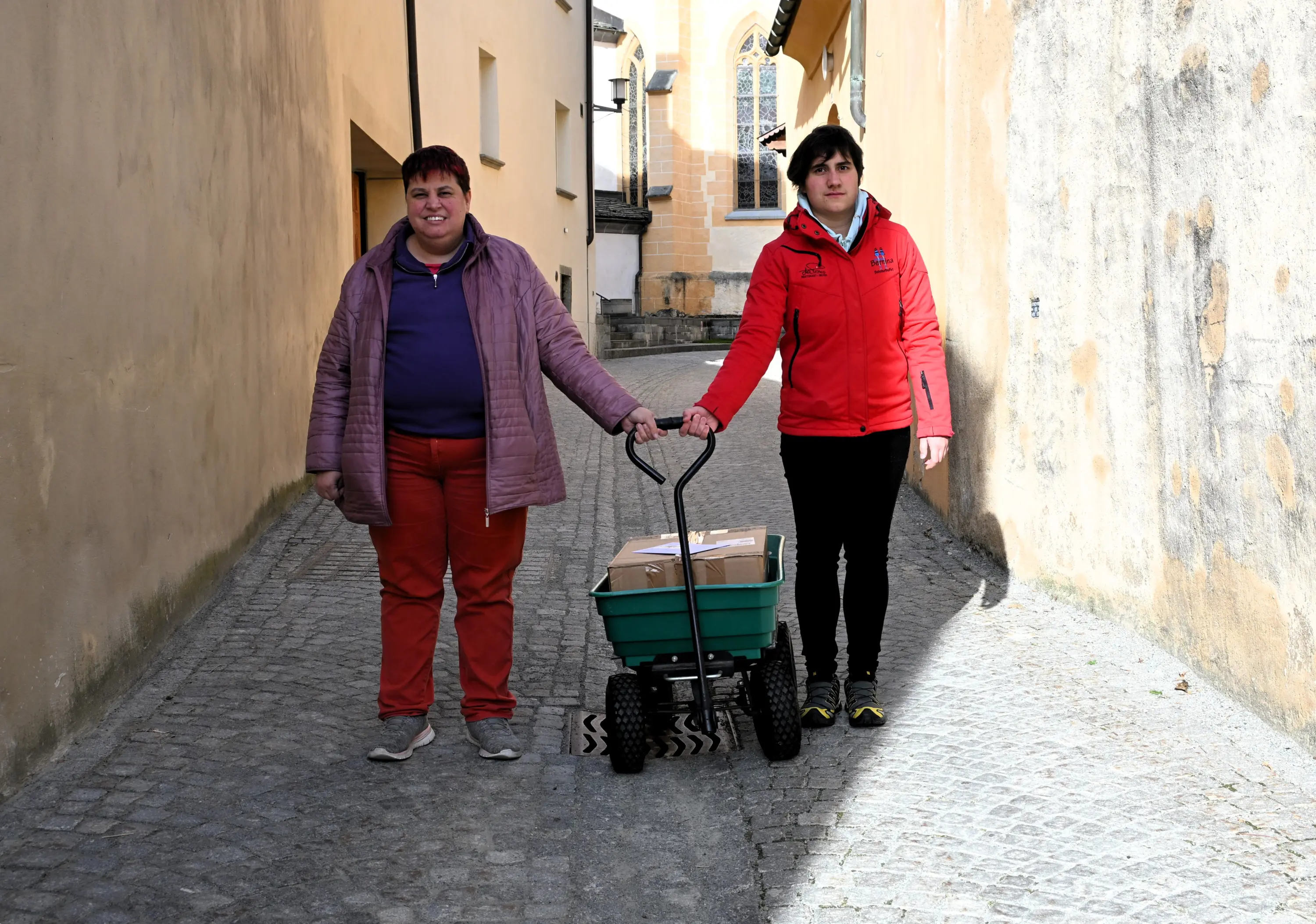Due persone in un vicolo stretto, una con giacca rosa e pantaloni rossi e l'altra con una giacca rossa. Stanno spingendo un carrello verde con pac