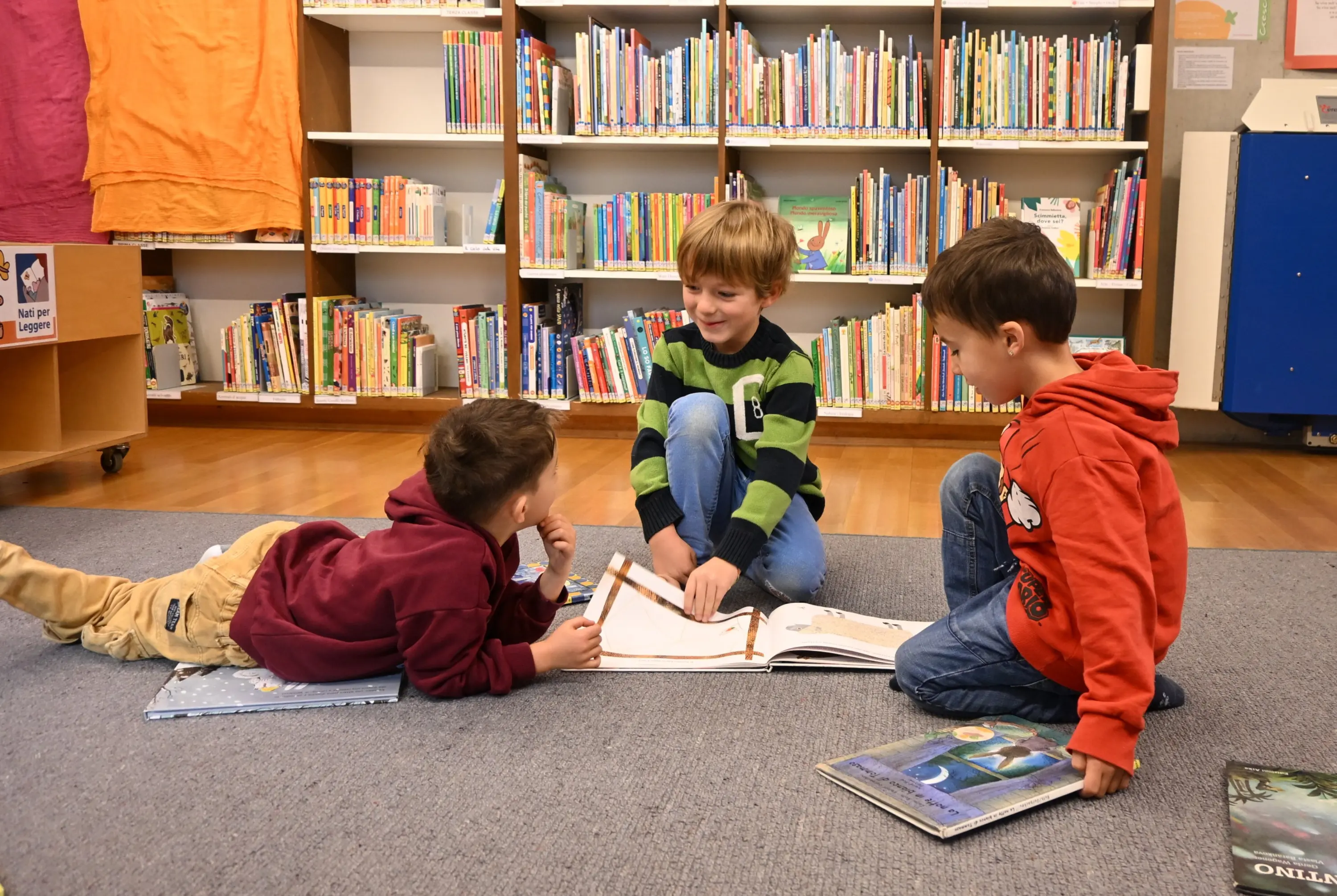 Tre bambini seduti sul pavimento di una biblioteca, intenti a sfogliare un libro illustrato. Due bambini sono sdraiati, mentre uno è seduto al centro. Sullo