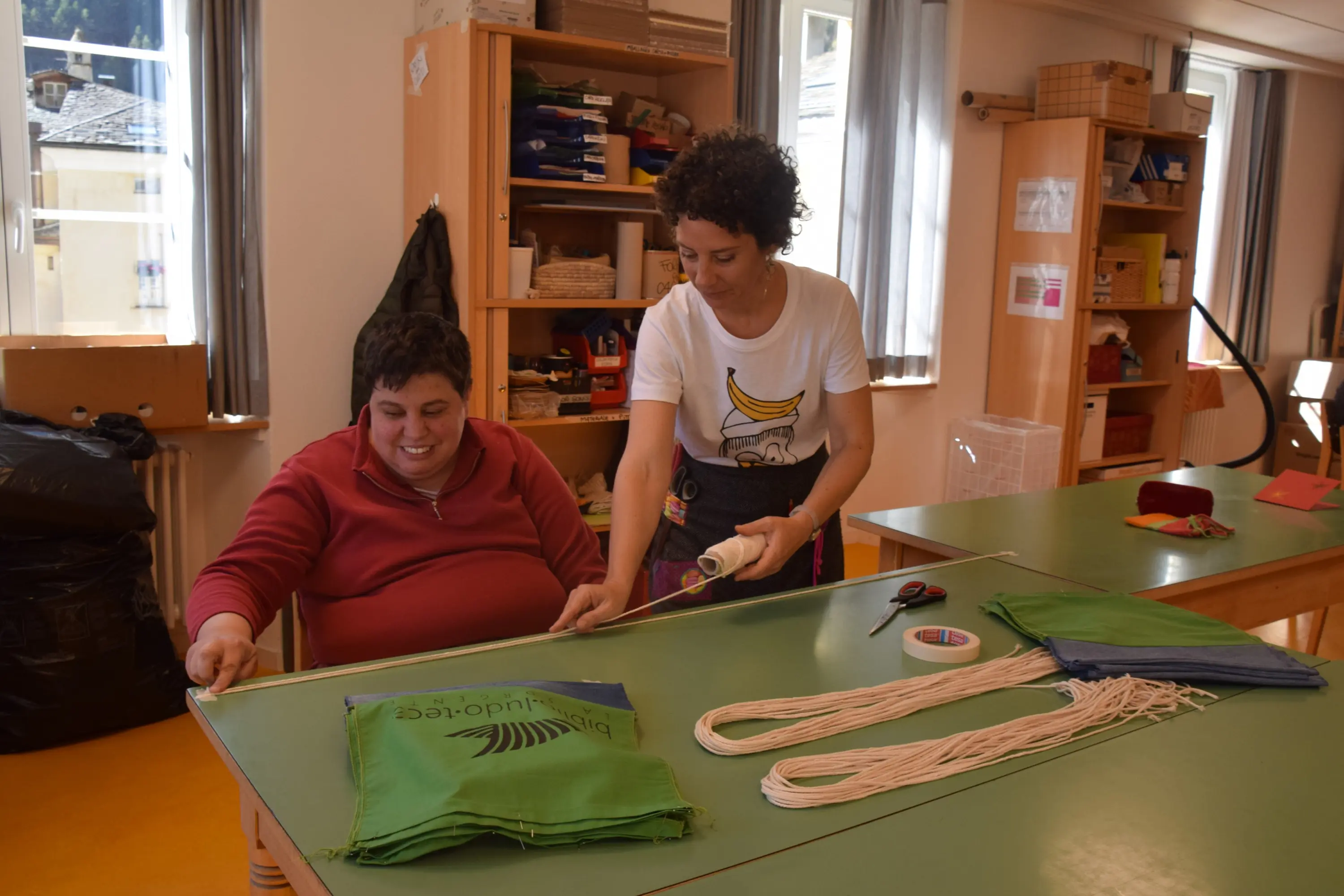 Due persone stanno lavorando insieme a un tavolo. Una donna in maglietta bianca con una banana disegnata sta aiutando un uomo a legare delle corde a sacchetti verdi