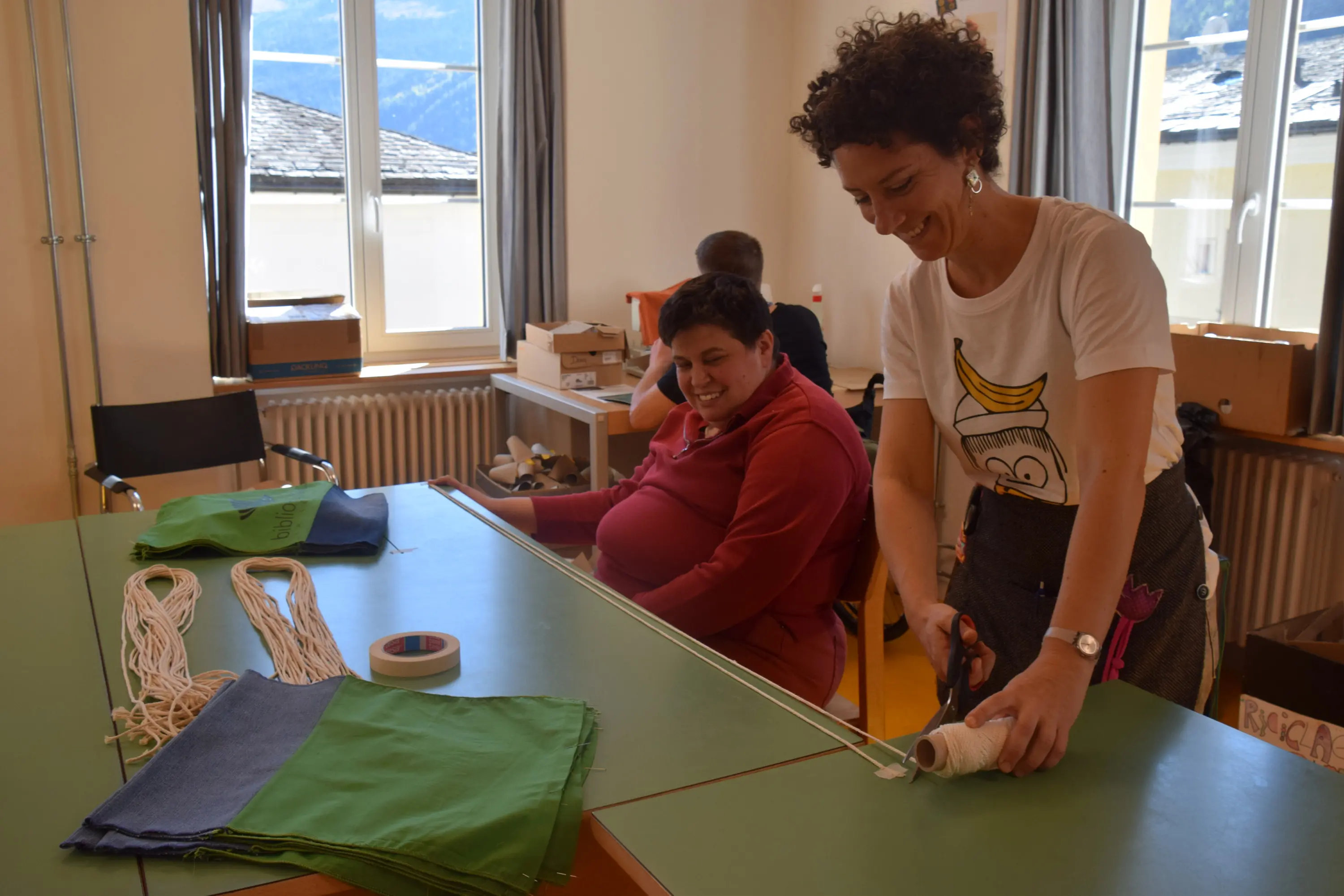Una donna con i capelli ricci taglia del filato mentre un uomo sorride seduto accanto a lei. Sul tavolo ci sono tessuti colorati e del nastro adesivo.