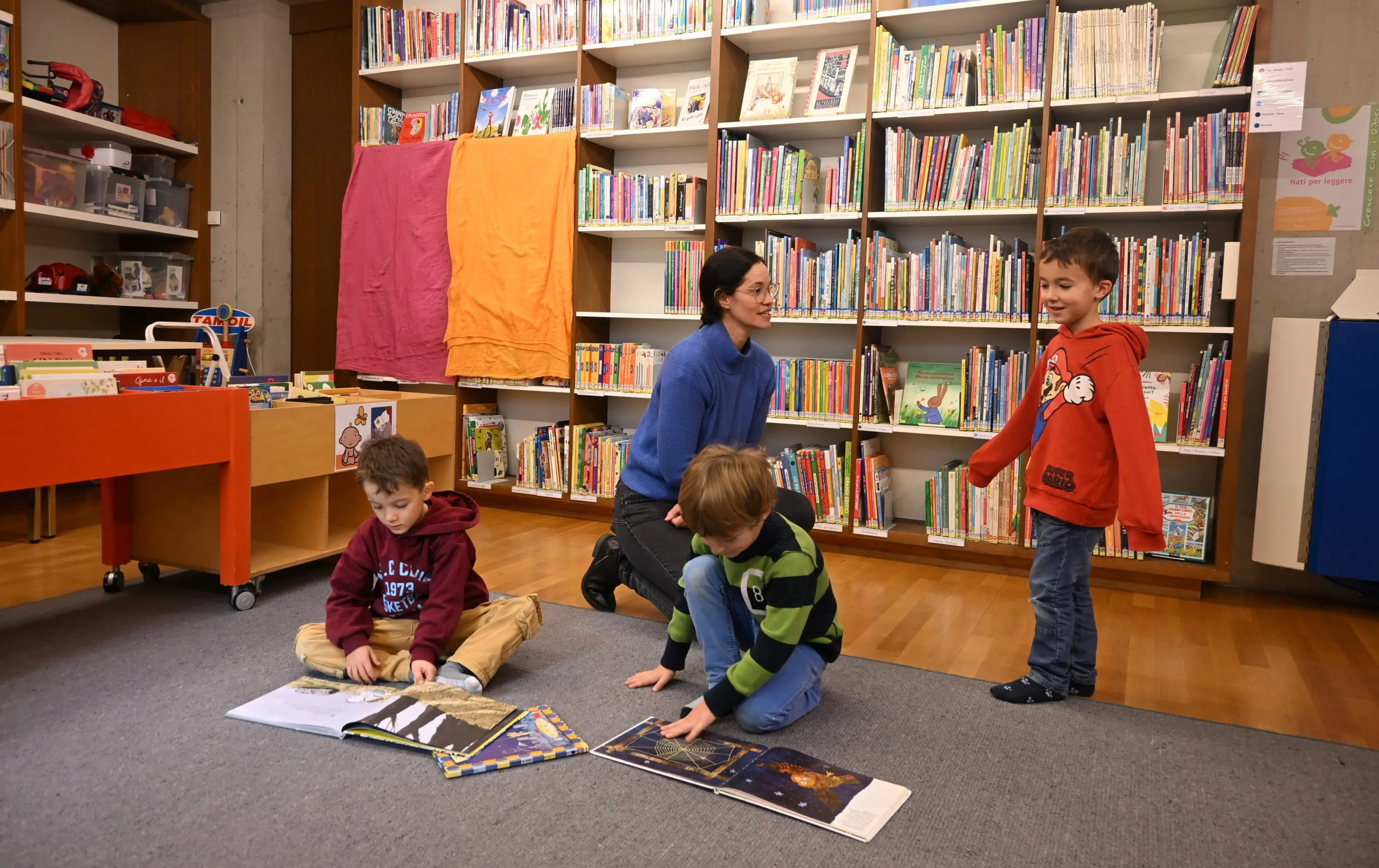 Un'insegnante interagisce con tre bambini in una biblioteca. Due bambini sono a terra mentre sfogliano dei libri illustrati, mentre il terzo bambino sta in piedi, sorrid