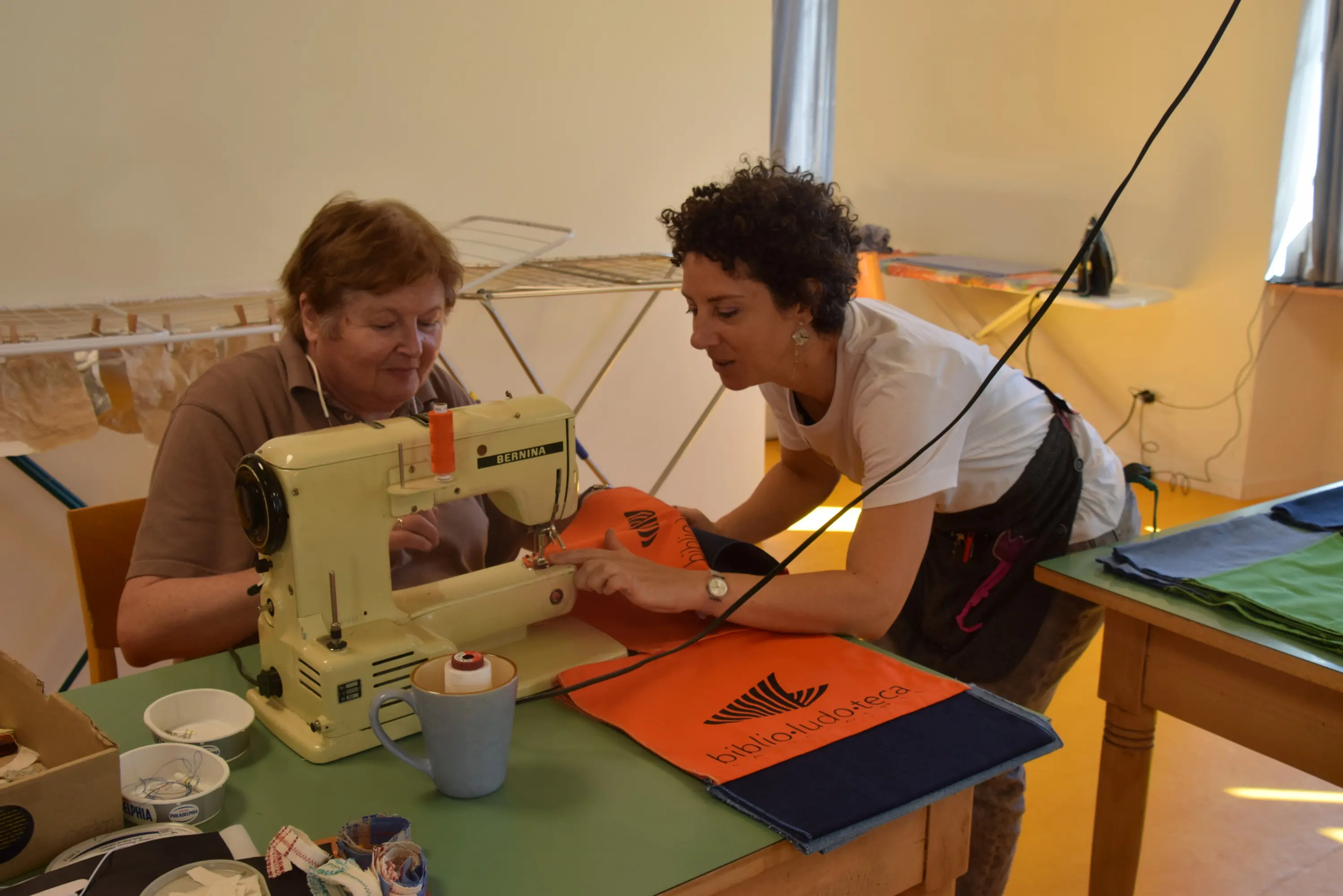 Due donne sono sedute a un tavolo, mentre una di loro guida l'altra nell'uso di una macchina da cucire. Sul tavolo ci sono alcune stoffe colorate e un pecc
