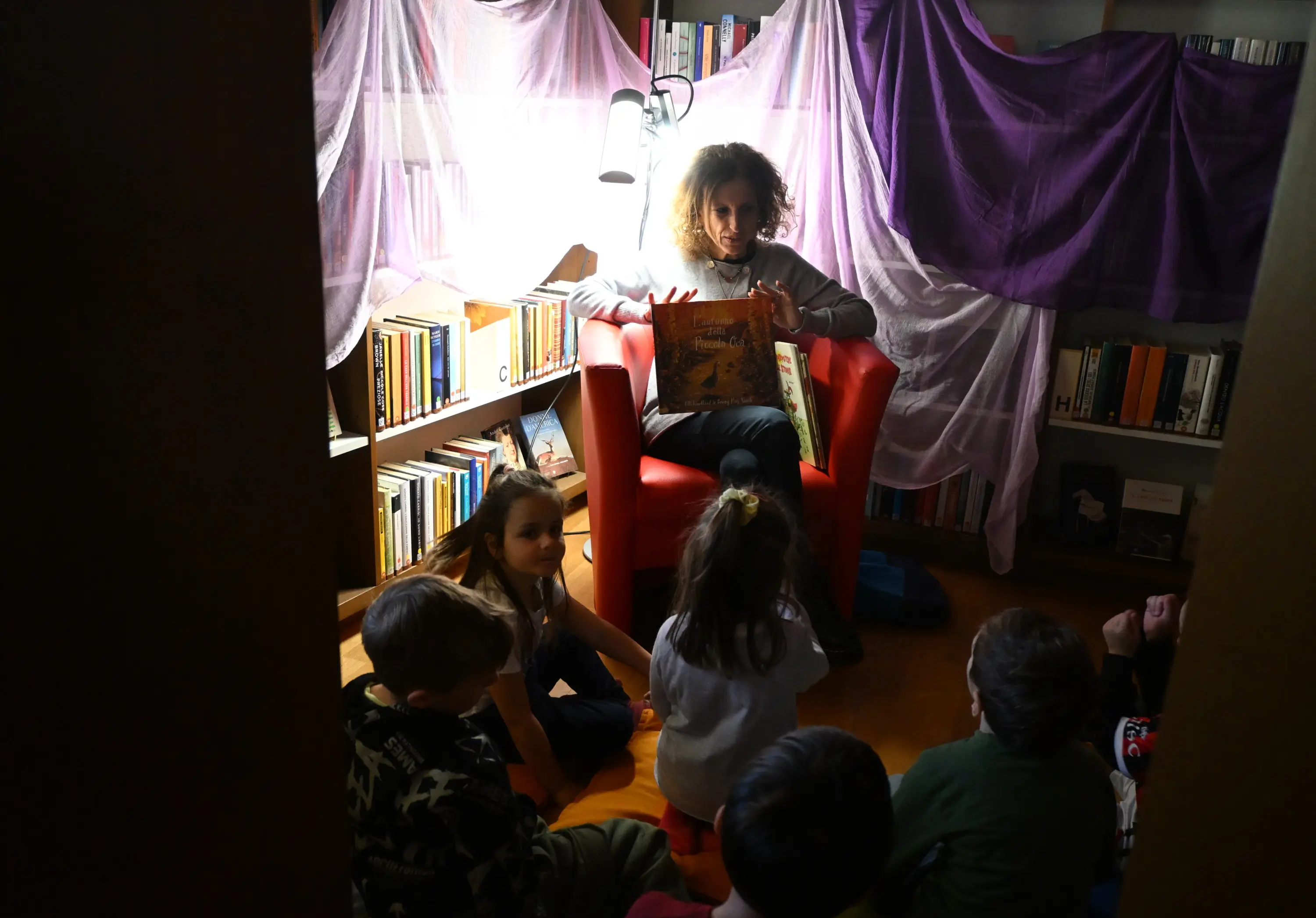 Un'educatrice legge un libro ai bambini seduti a terra, circondati da scaffali di libri e teli colorati che creano un'atmosfera accogliente.