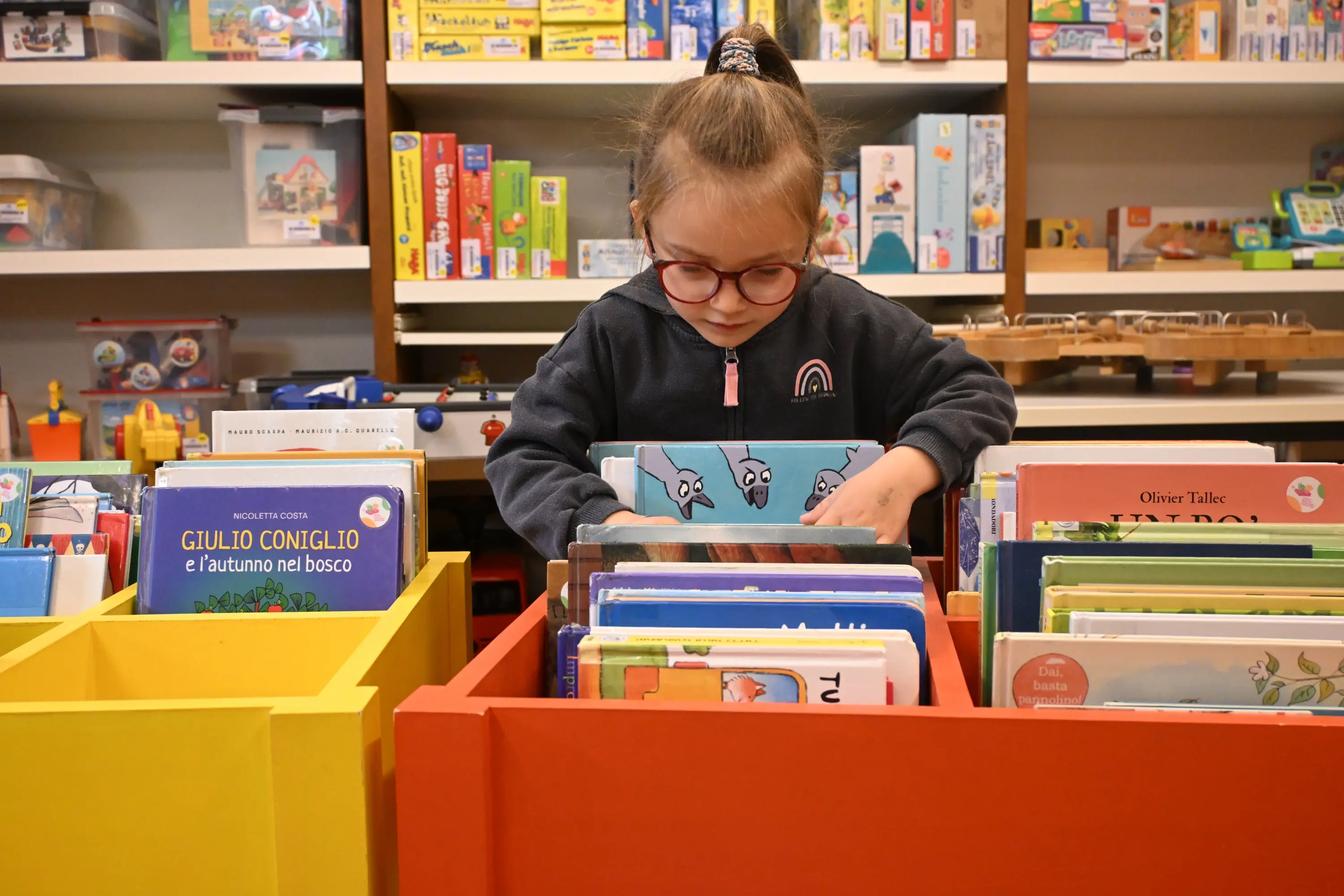 Una bambina con occhiali sta sfogliando dei libri in una libreria colorata con cassetti gialli e rossi. Sullo sfondo ci sono giochi e altri libri