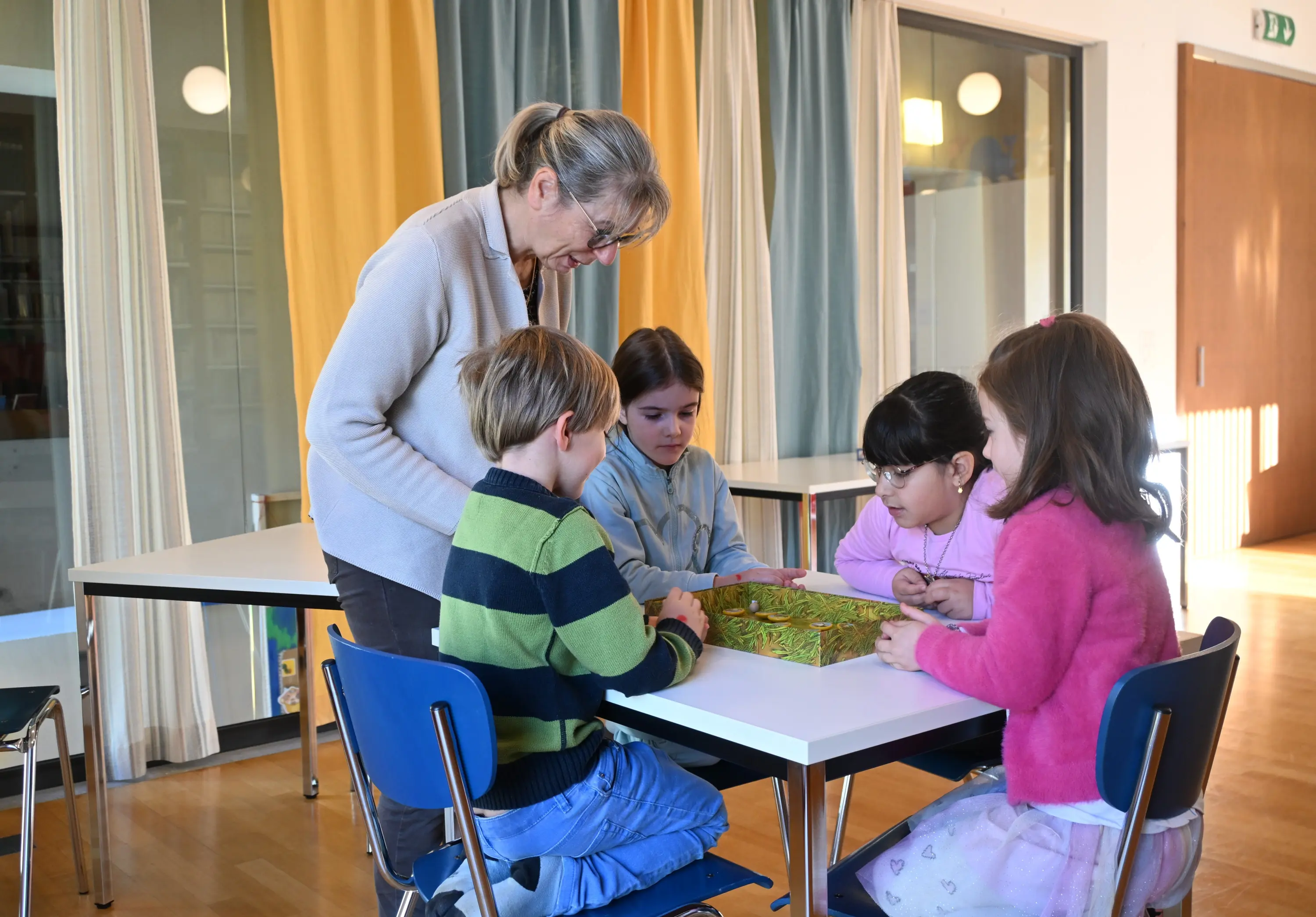 Una donna anziana supervisa un gruppo di quattro bambini mentre giocano a un gioco da tavolo in una stanza luminosa con tende colorate.