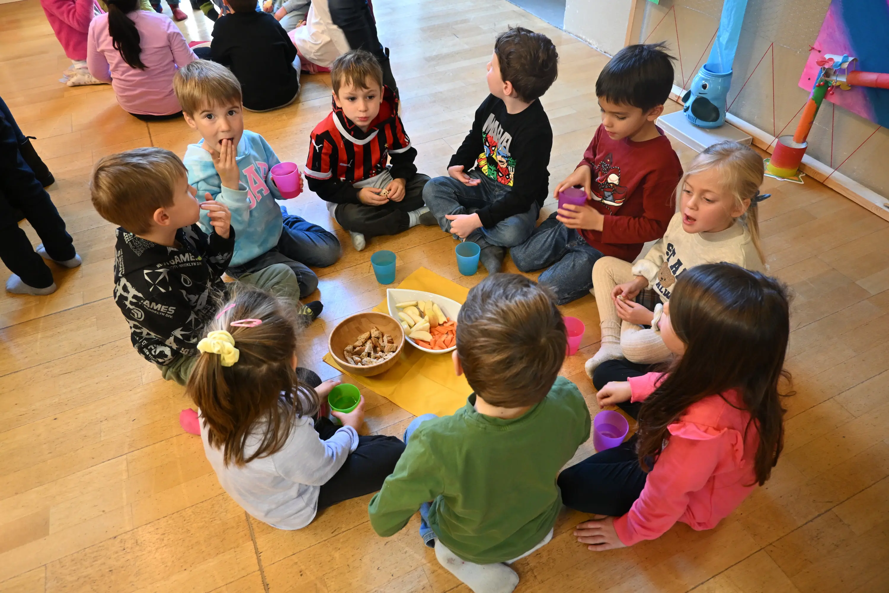 Un gruppo di bambini seduti a terra in cerchio. Stanno condividendo cibo e bevande, con piatti di snack al centro. Alcuni bambini stanno bevendo da bicch