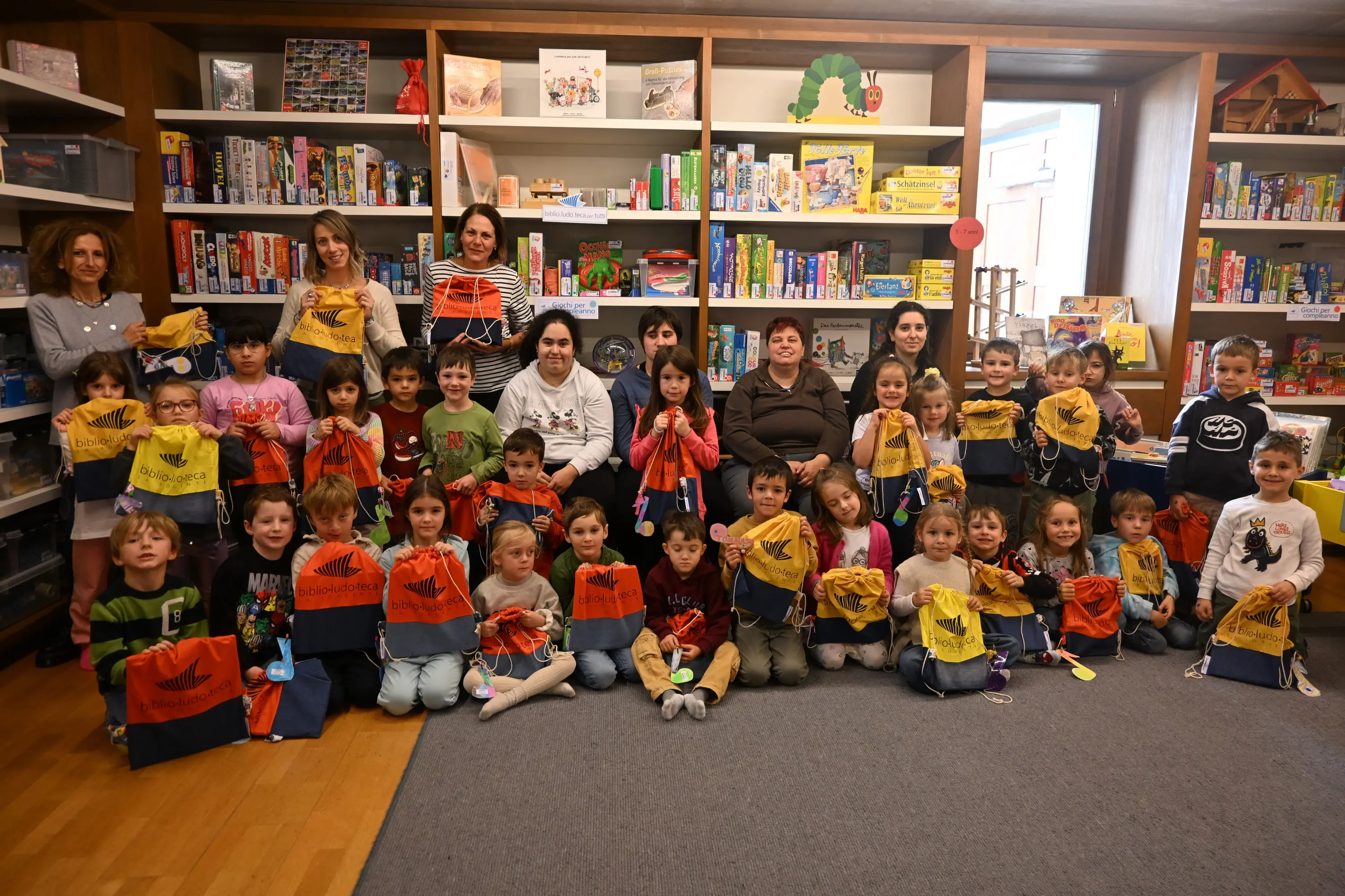 Gruppo di bambini e adulti sorridenti in un ambiente domestico, con scaffali pieni di giochi e libri sullo sfondo. I bambini tengono in mano dei sacchetti colorati
