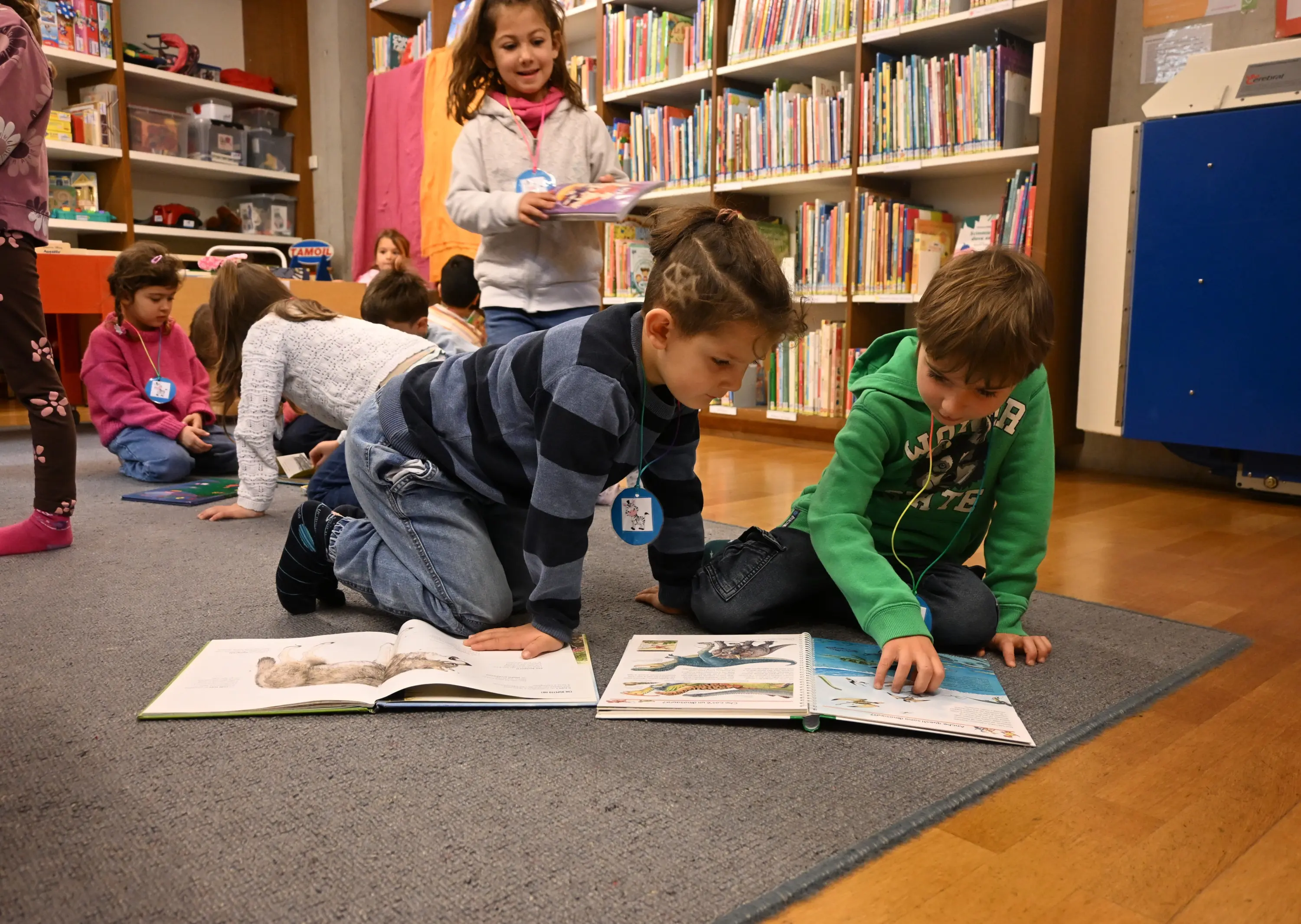 Bambini che leggono libri a un tavolo in una biblioteca, alcuni seduti a terra, altri in piedi, immersi nella lettura e nell'esplorazione dei testi.
