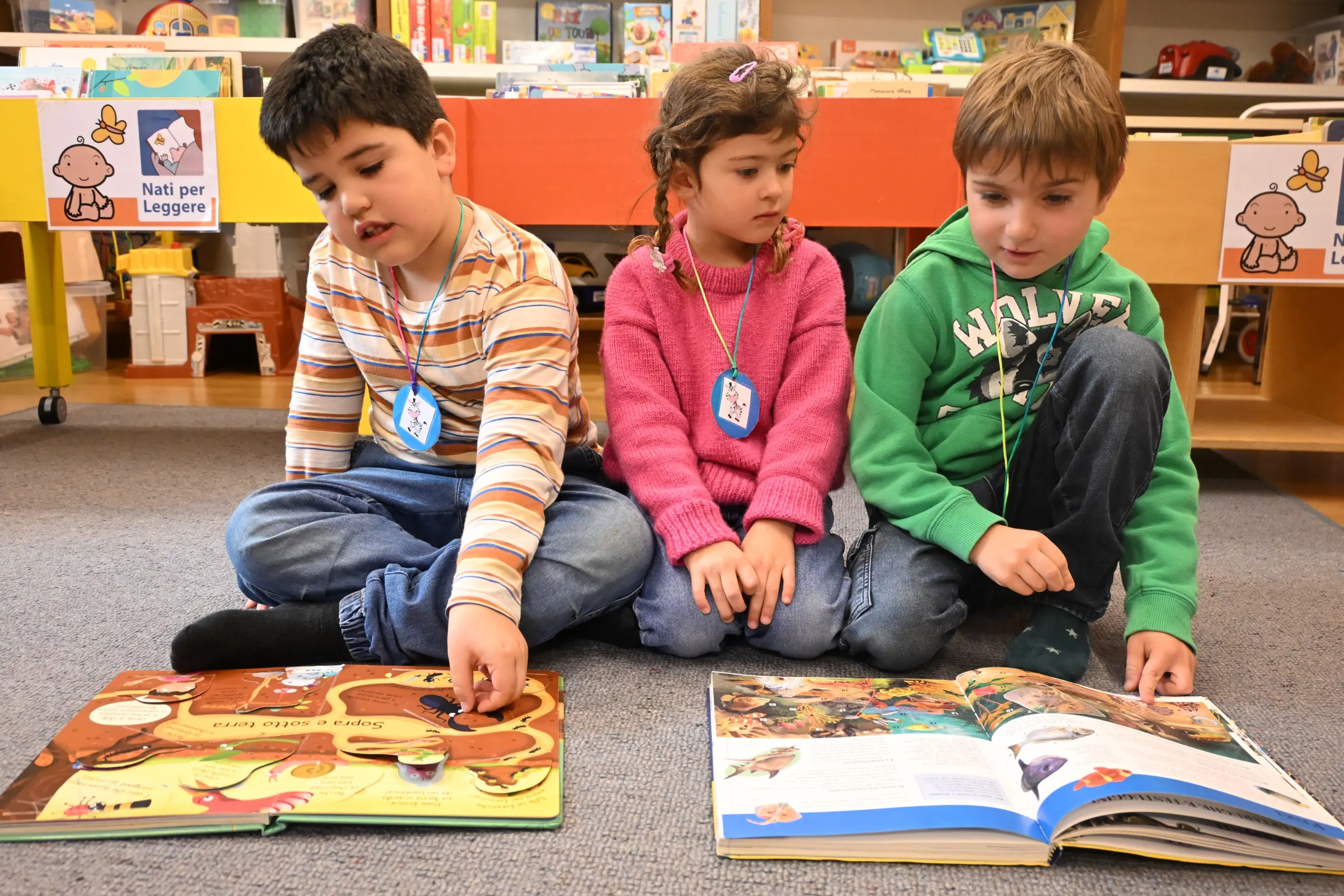 Tre bambini seduti a terra in una biblioteca, intenti a leggere libri illustrati. Due bambini, un ragazzo e una ragazza, sono a sinistra, mentre un altro ragazzo è