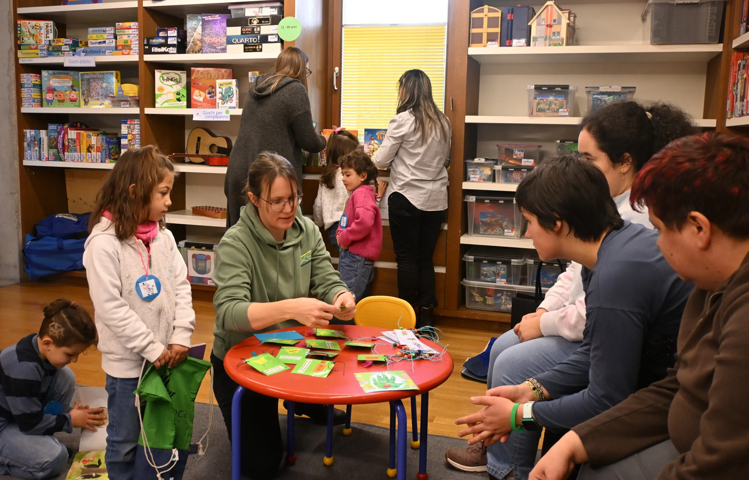 Una donna spiega un'attività a un gruppo di bambini e adulti seduti attorno a un tavolo colorato in una biblioteca. Sul tavolo ci sono materiali di artigianato