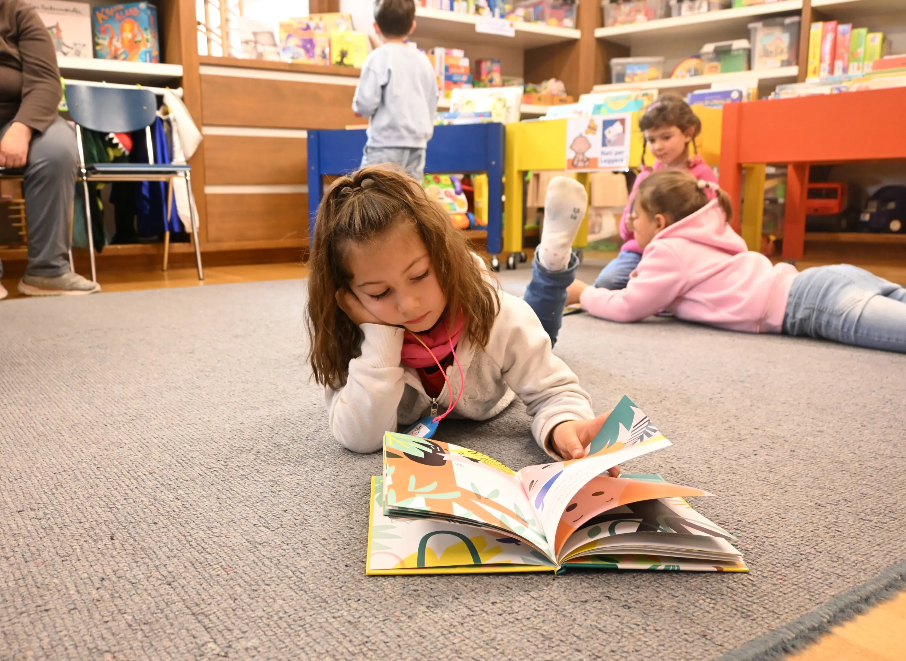 Un gruppo di bambini in una biblioteca. Una bambina è distesa sul pavimento, intenta a leggere un libro illustrato, mentre altri bambini giocano o leggono nei pressi.