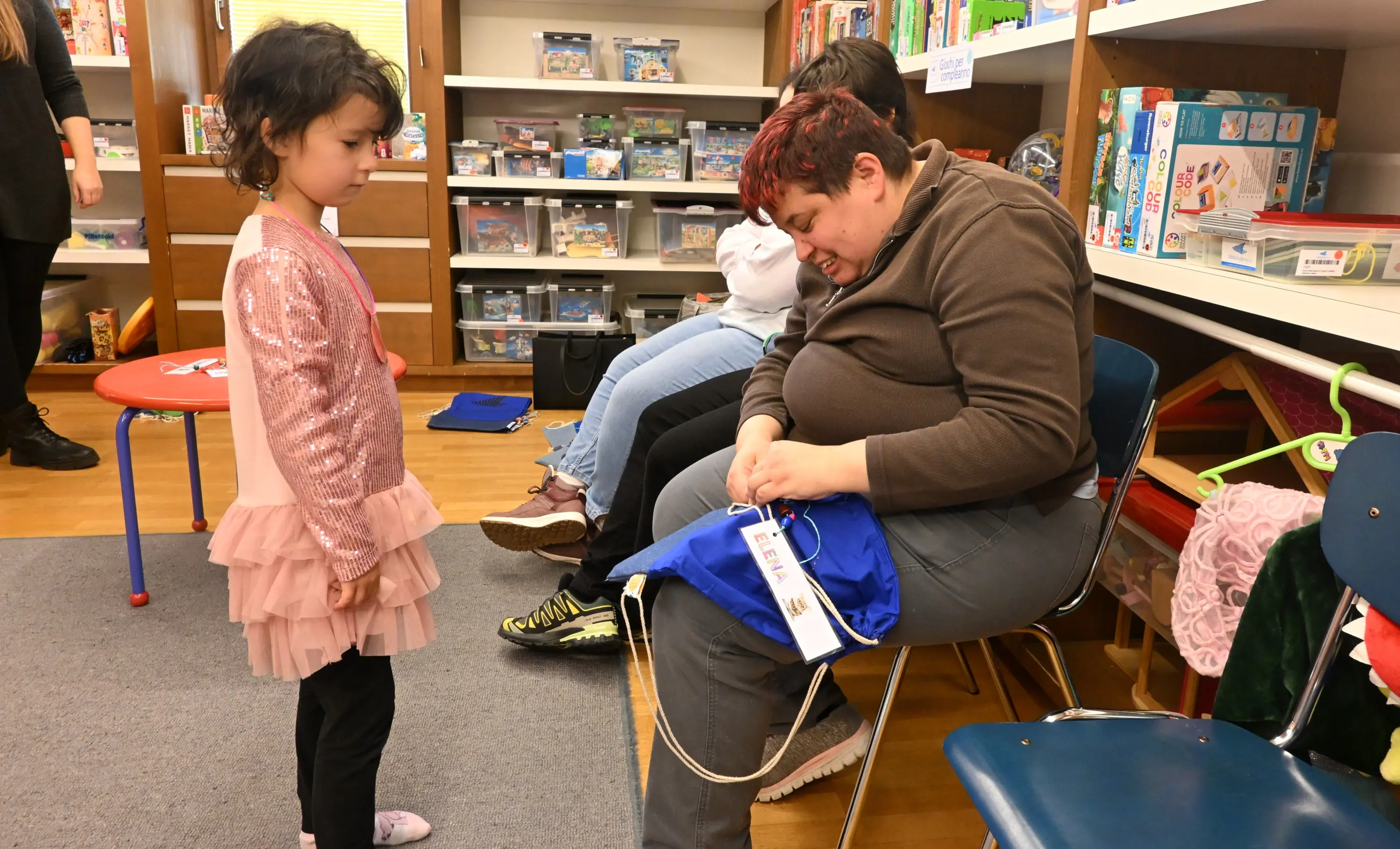Un bambino in abito rosa osserva attentamente un adulto seduto che sta preparando un sacchetto blu, circondato da giocattoli e libri. Altre persone sono sedute sul